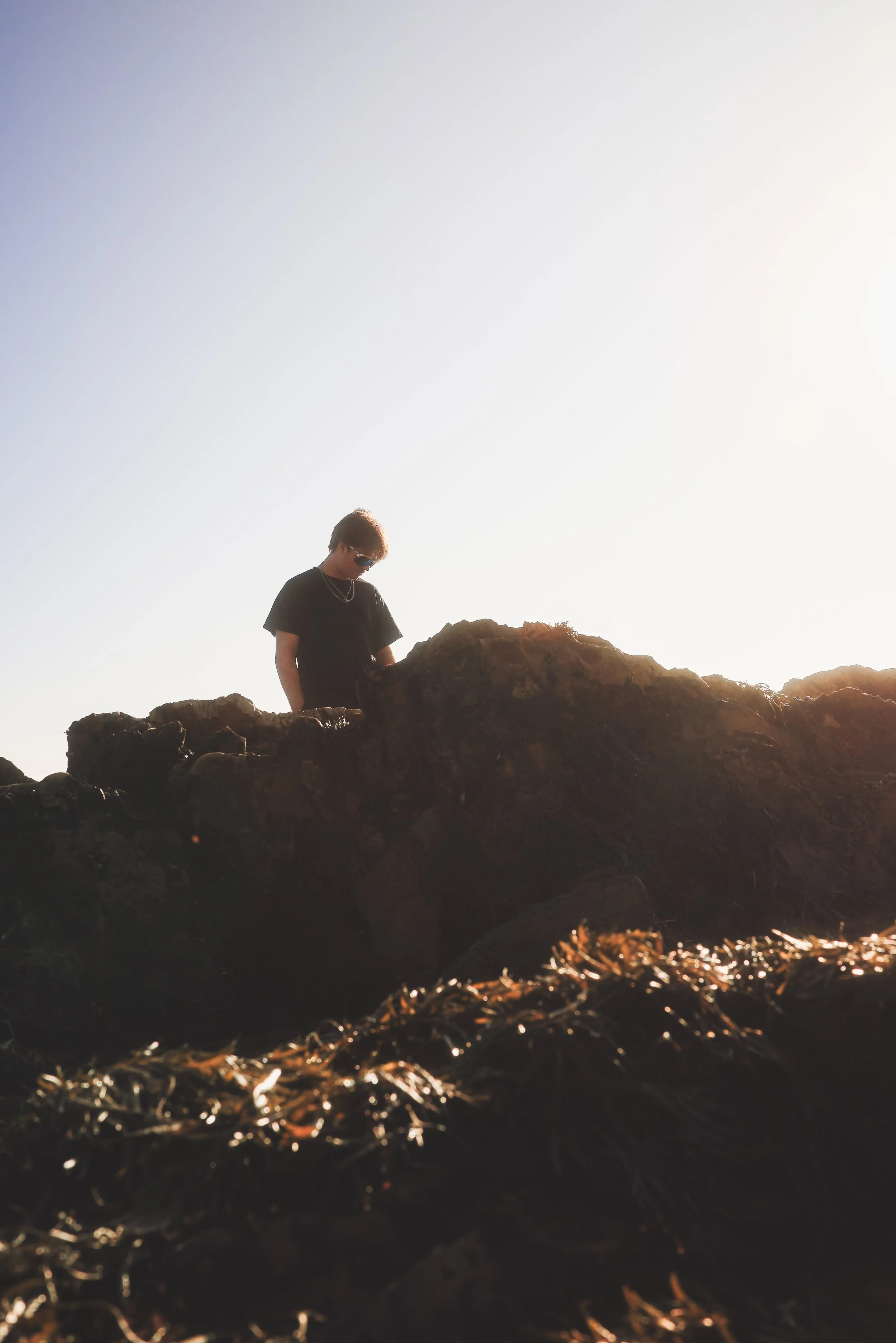 A person wearing sunglasses and a black shirt standing on rocks at sunset or sunrise.