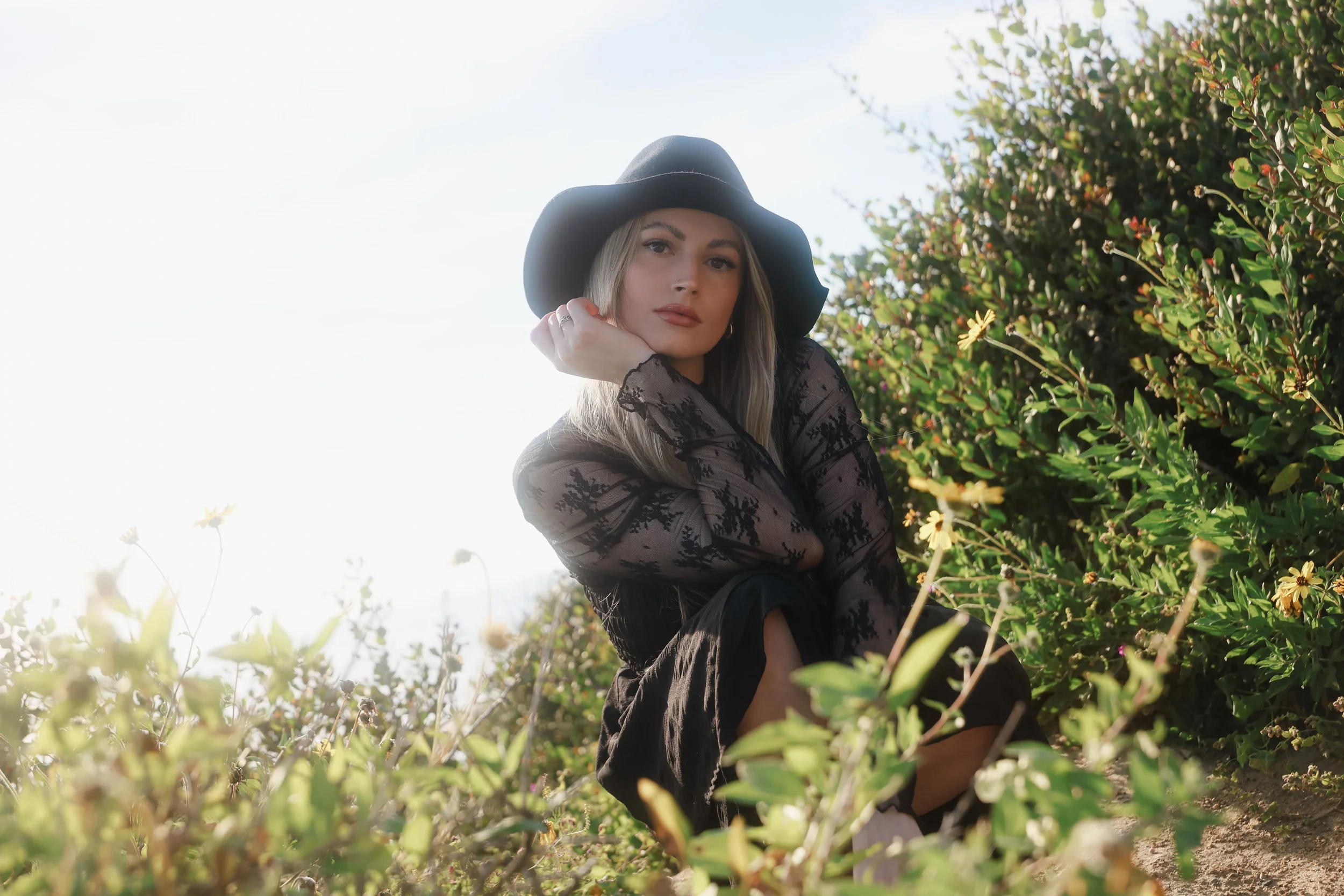 Young woman in a black dress and wide-brimmed hat sitting among green shrubs on a sunny day.