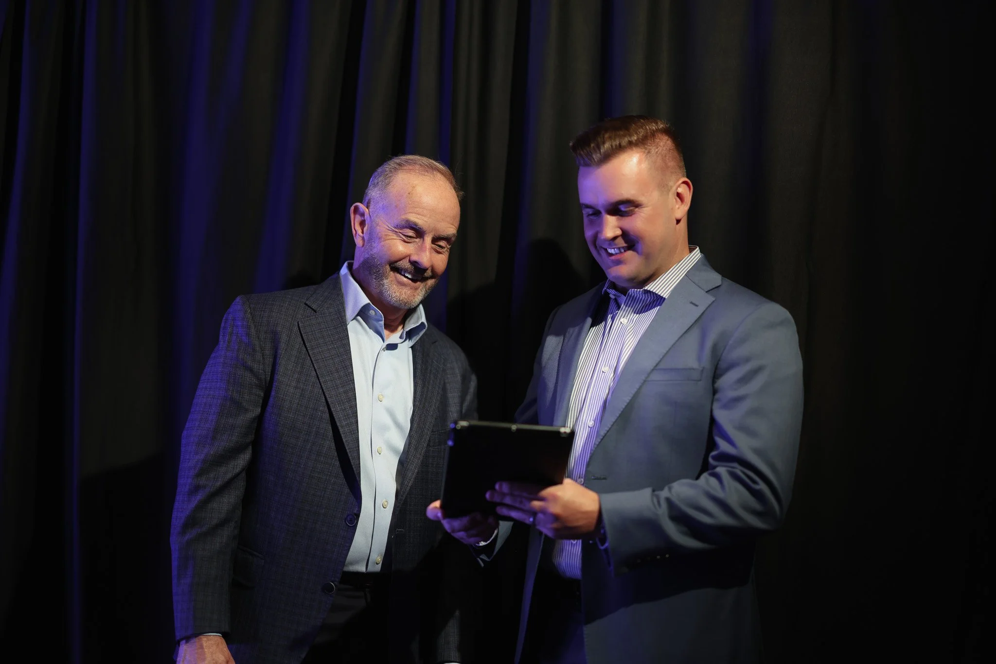 Two men in suits are smiling and looking at a tablet, standing in front of black curtains.