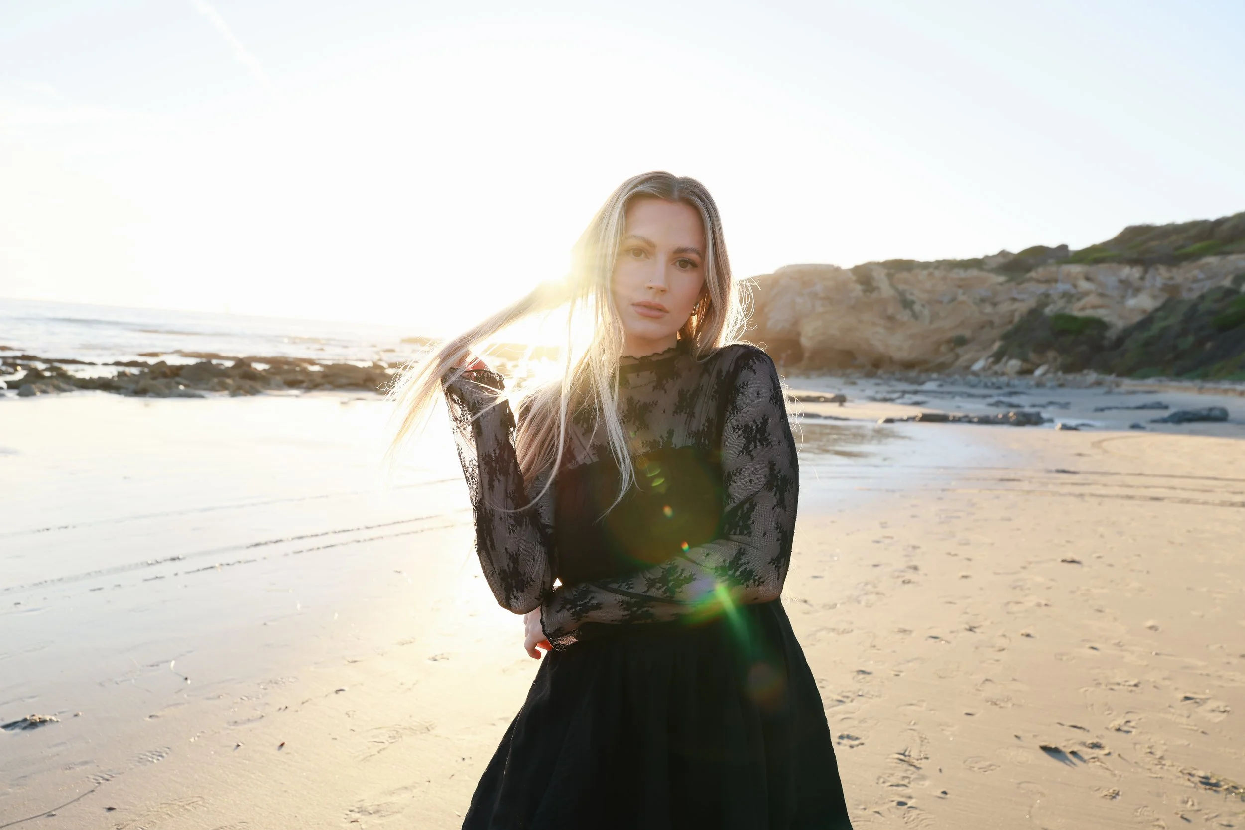 A woman with long blonde hair in a black lace dress stands on a sandy beach during sunset, with cliffs in the background and sunlight creating a glow around her.