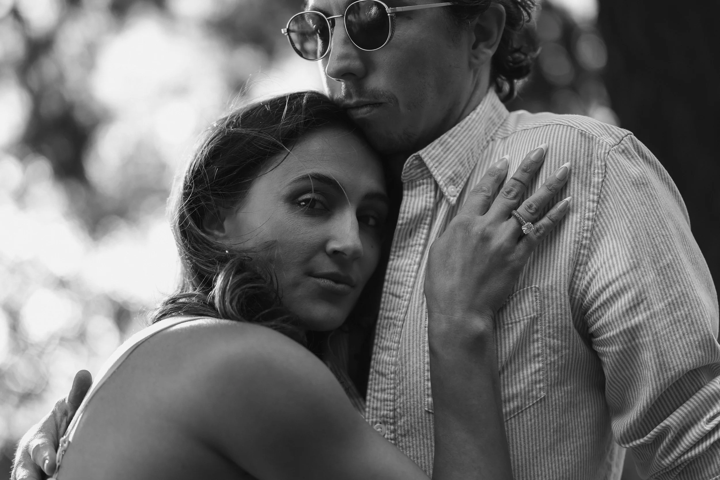 A black and white photo of a man and woman embracing closely. The woman rests her head on the man's chest, looking at the camera, while the man wears sunglasses and touches her shoulder gently.