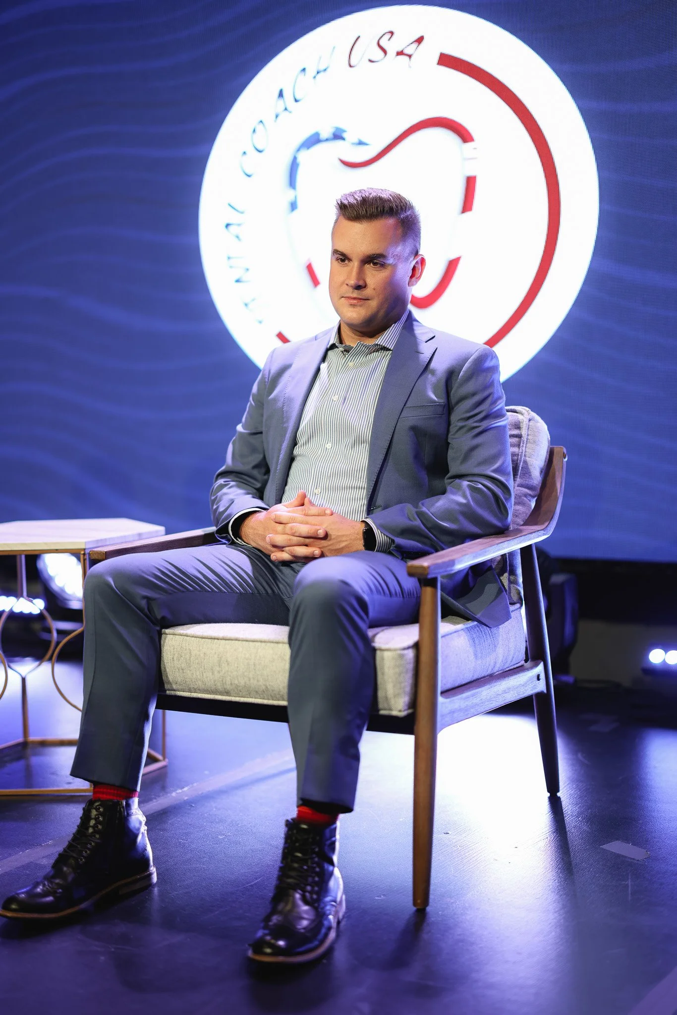 A man in a gray suit and striped shirt is sitting on a beige armchair with wooden arms, on a stage with a dark background. Behind him is a large, round logo with a blue and red design and text that appears to spell 'NATOACH USA.' The stage has some l