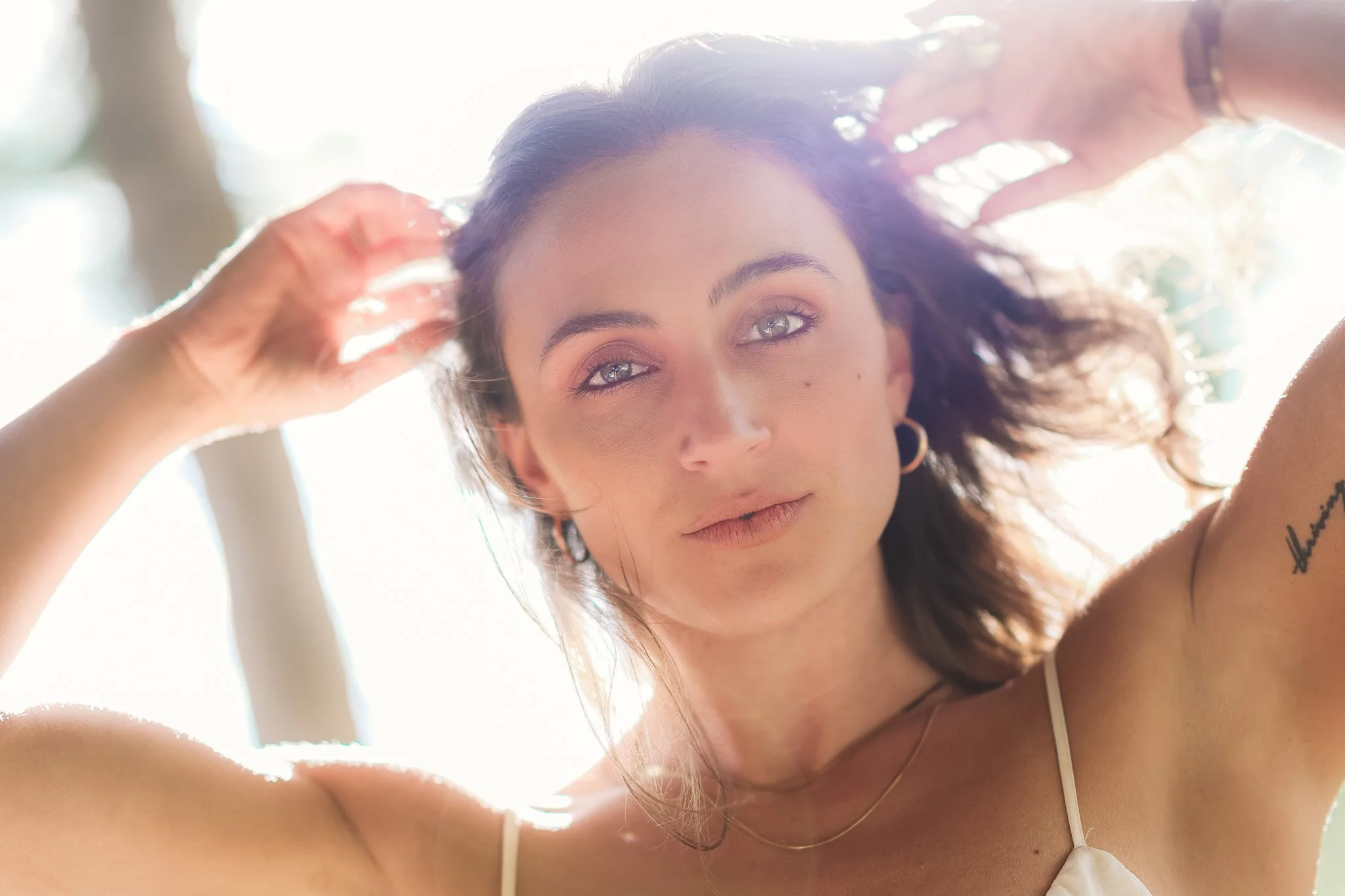 A woman with curly hair and gray eyes looks into the camera, with sunlight shining behind her and some hands adjusting her hair.