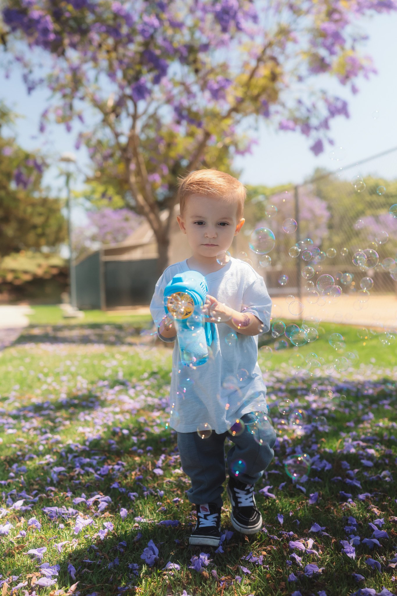 A young boy with red hair and a white shirt walking through a park covered with purple flowering petals. He is holding a bubble gun, and bubbles are floating around him. The background features a purple-flowering tree and a fence.