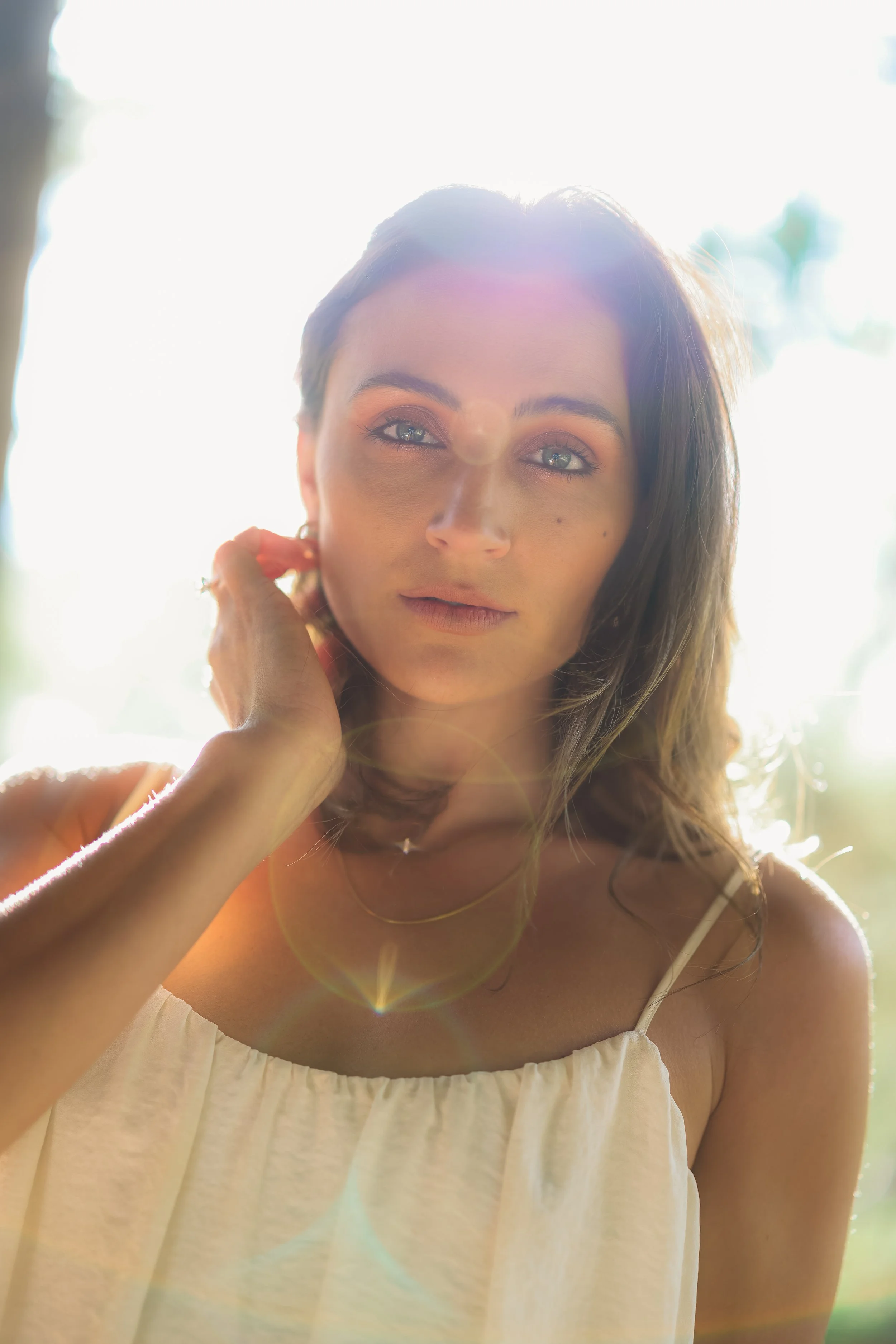 A woman smiling outdoors in bright sunlight, wearing a white spaghetti strap top and gold jewelry.