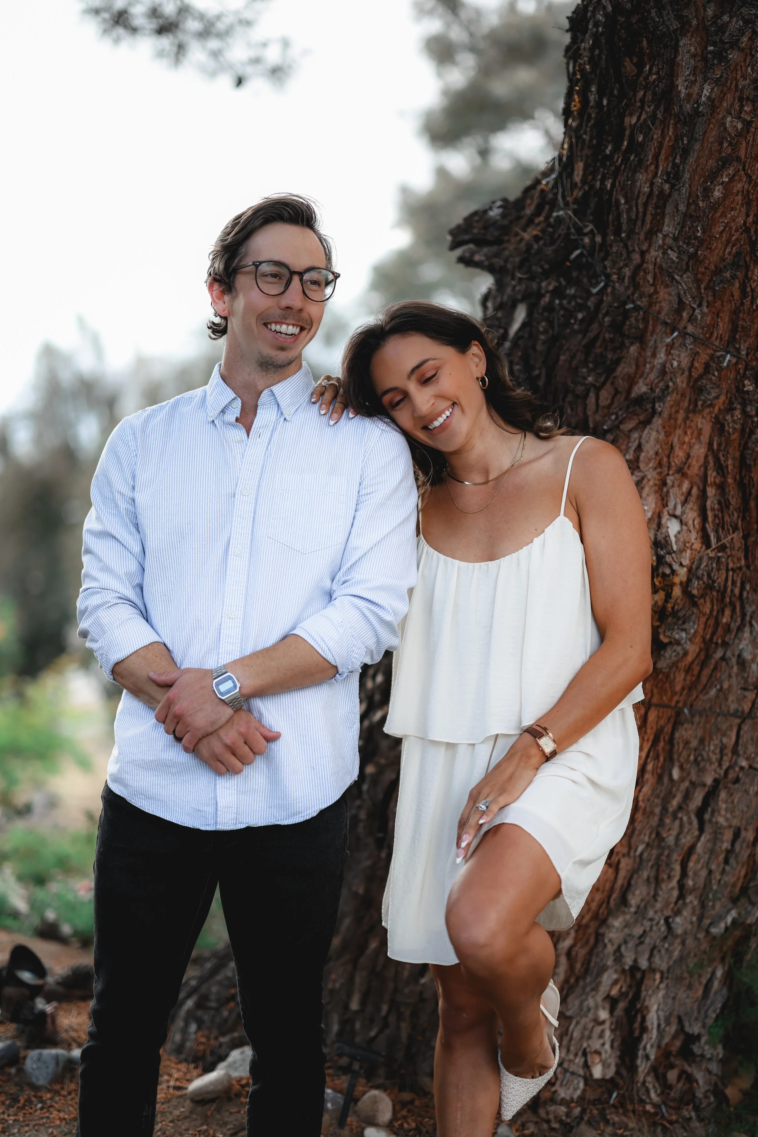A smiling man and woman standing next to a large tree, with the woman leaning her head on the man's shoulder.