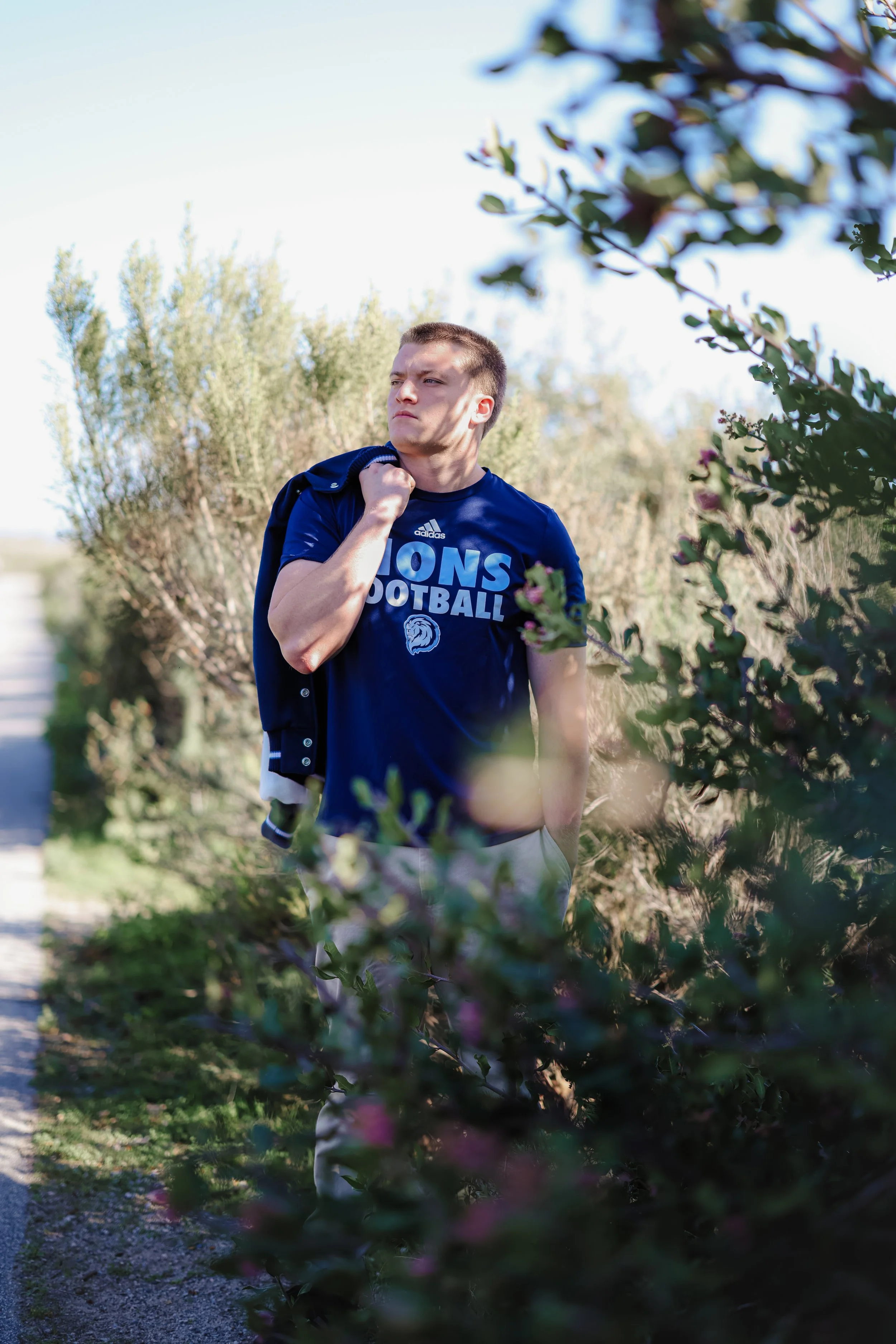 A young man in a blue football T-shirt stands outdoors among bushes, holding a jacket over his shoulder and looking pensively into the distance.
