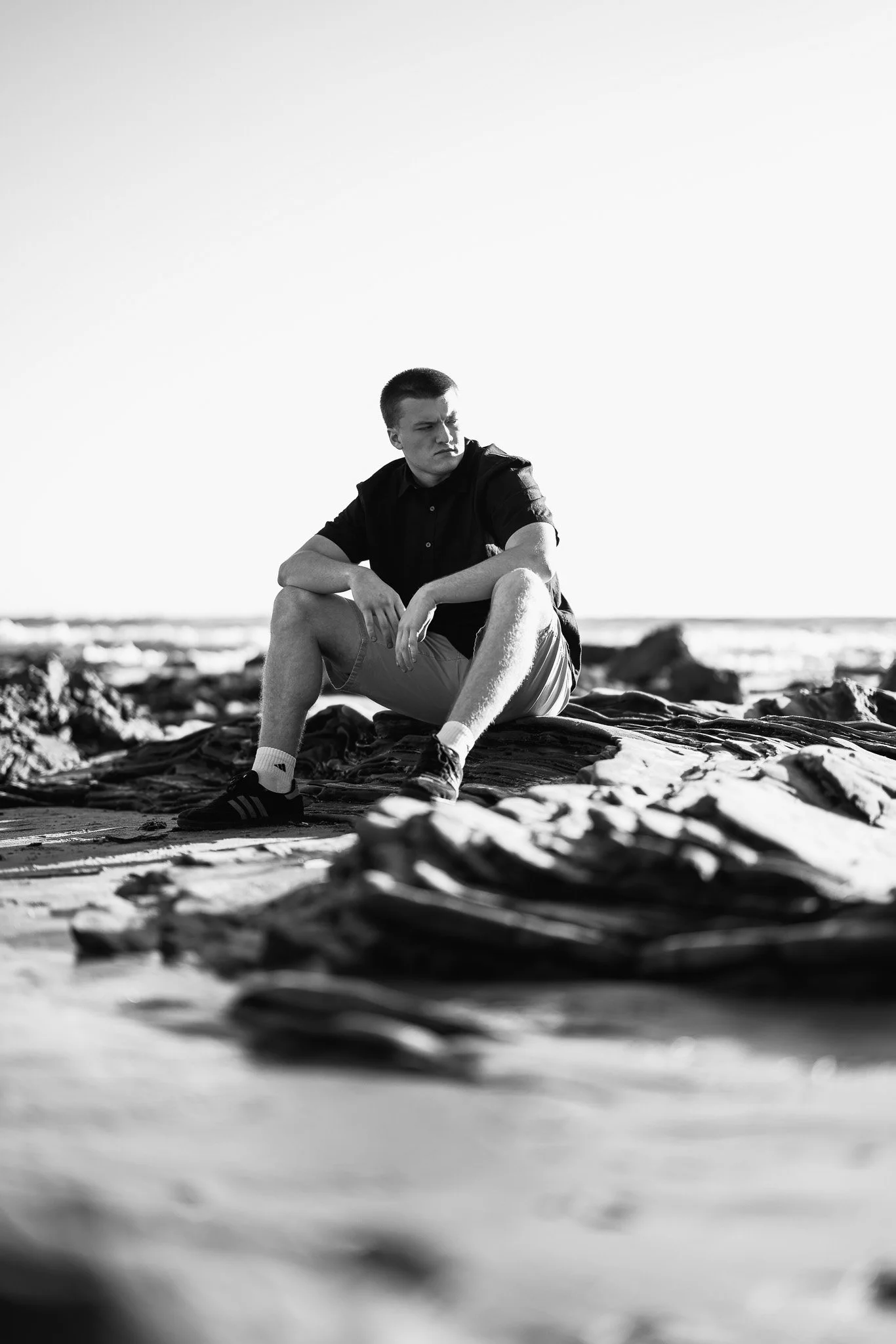 Black and white photo of a young man sitting on the beach on a large piece of driftwood or rock, looking to the side with a serious expression, with rocks and the ocean in the background.