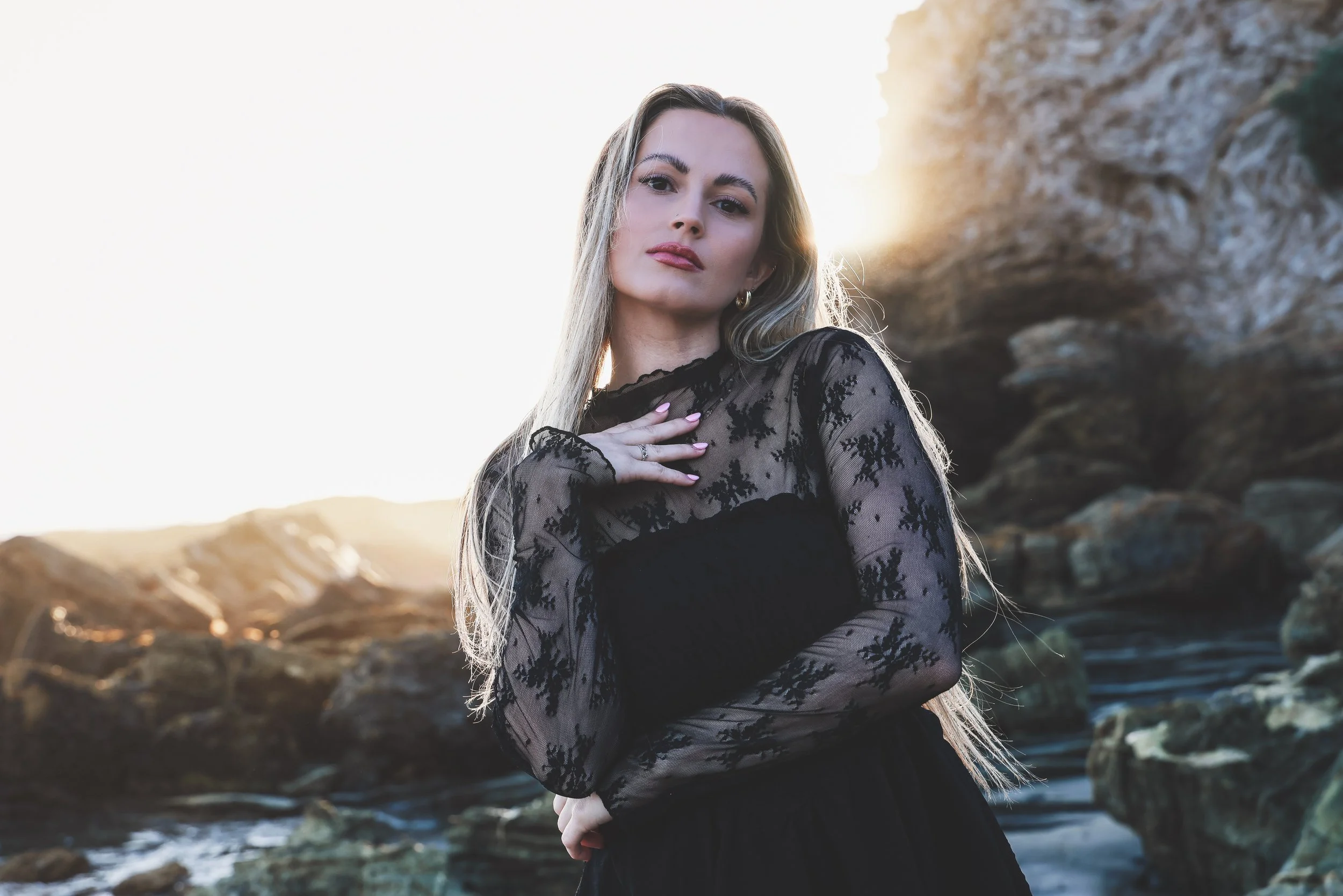 A young woman with blonde hair and fair skin standing outdoors during sunset, wearing a black lace dress, with her hand on her chest, near a rocky landscape.