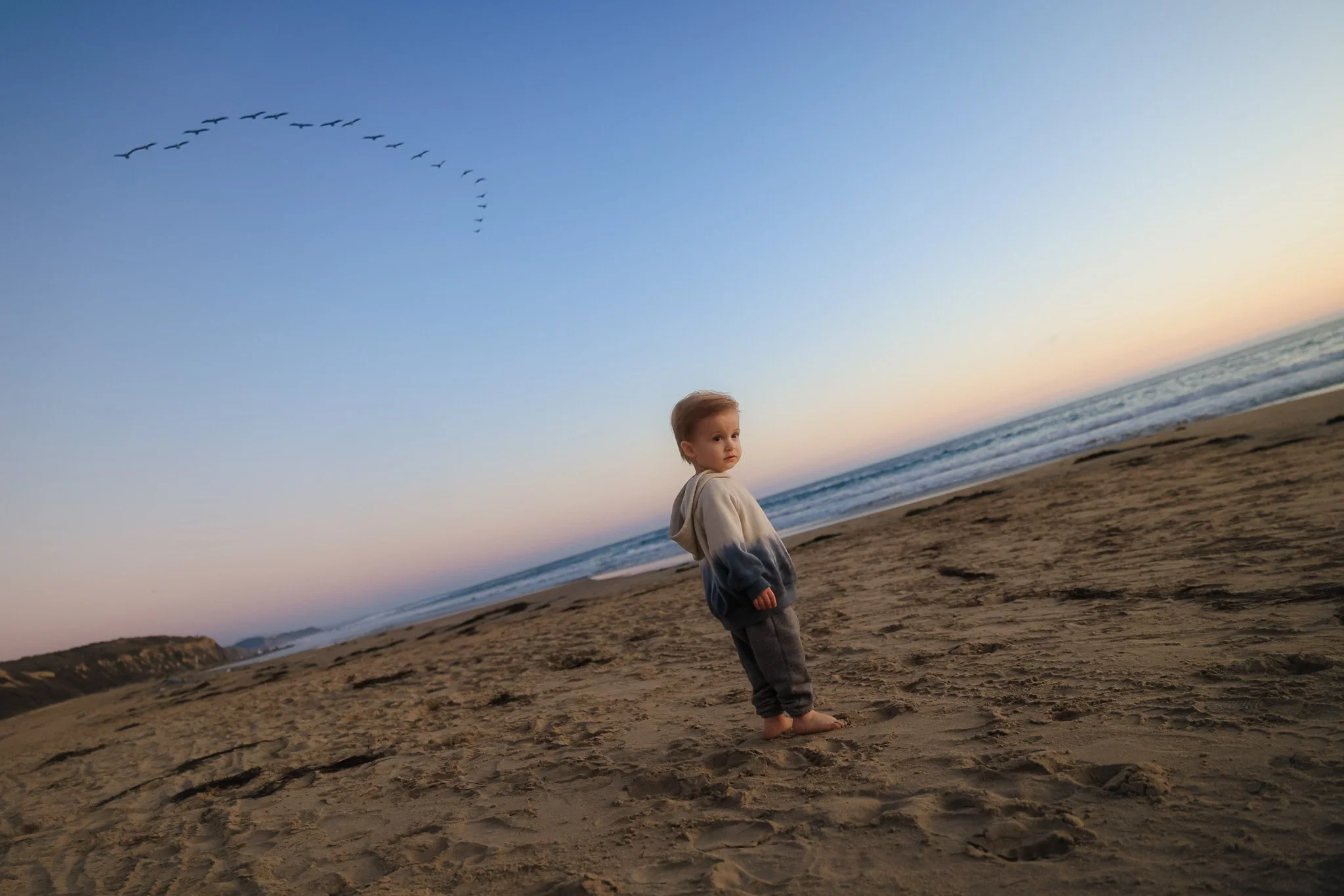 A young boy standing barefoot on a sandy beach during sunset, looking to the side with a cloudy sky and a flock of birds flying across.