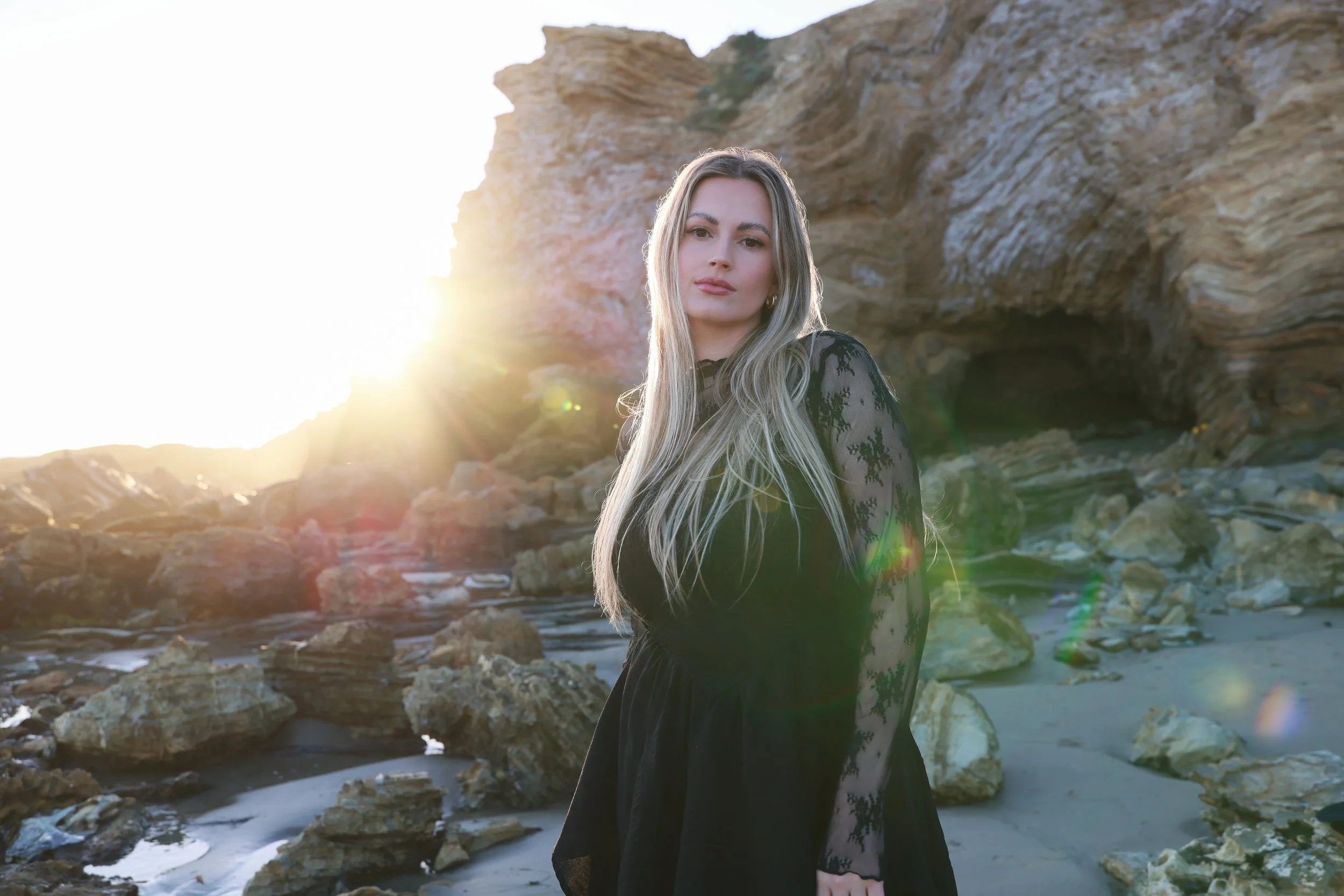A woman with long blonde hair in a black lace dress standing on a rocky beach at sunset with cliffs in the background.