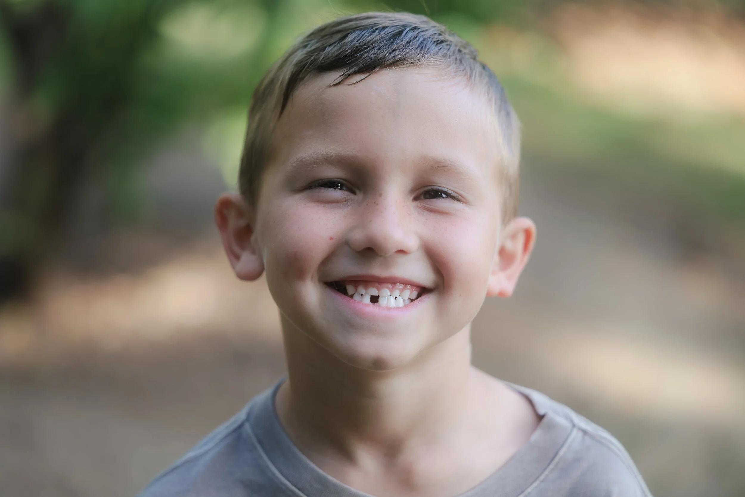 A young boy with short light brown hair and light skin, smiling outdoors with a blurred background of greenery.