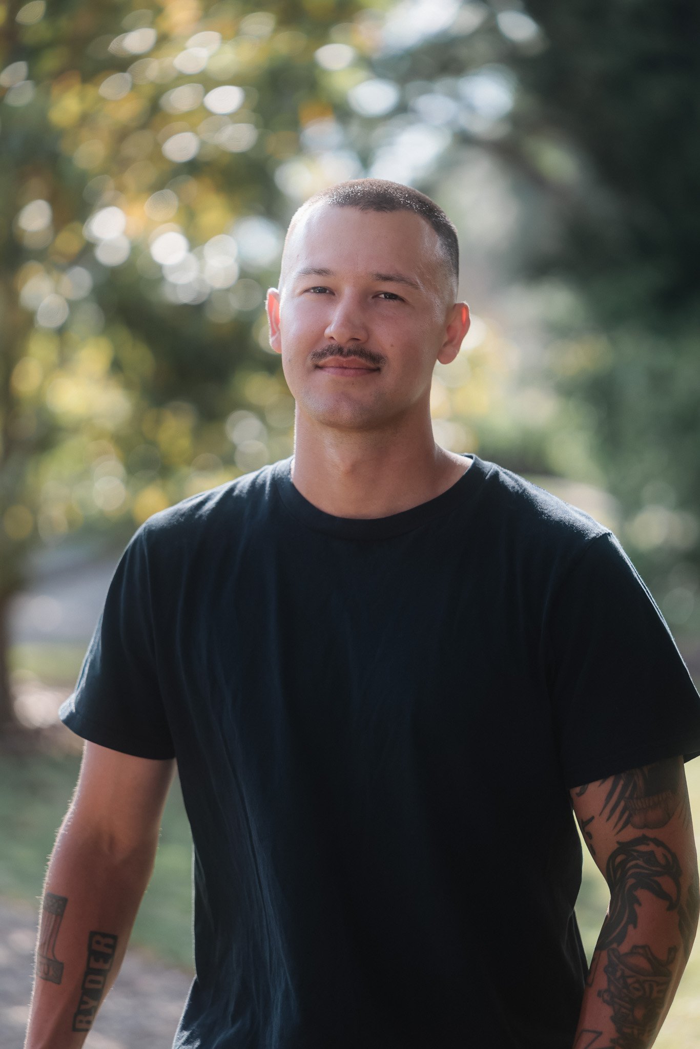 A man with short hair and a mustache, wearing a black T-shirt, standing outdoors with blurred trees and sunlight in the background.
