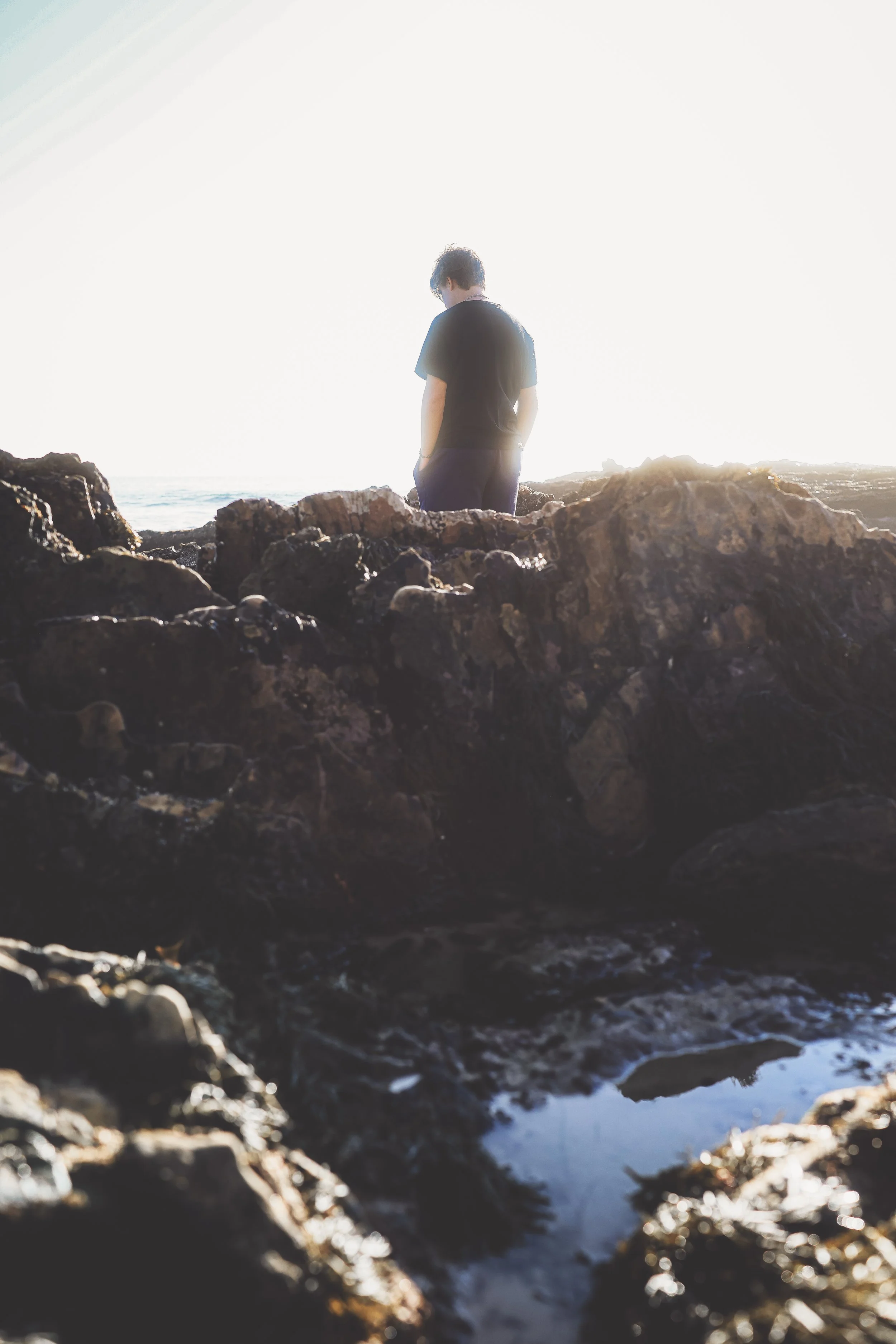 A person standing on rocks by the ocean during sunset, facing away from the camera.