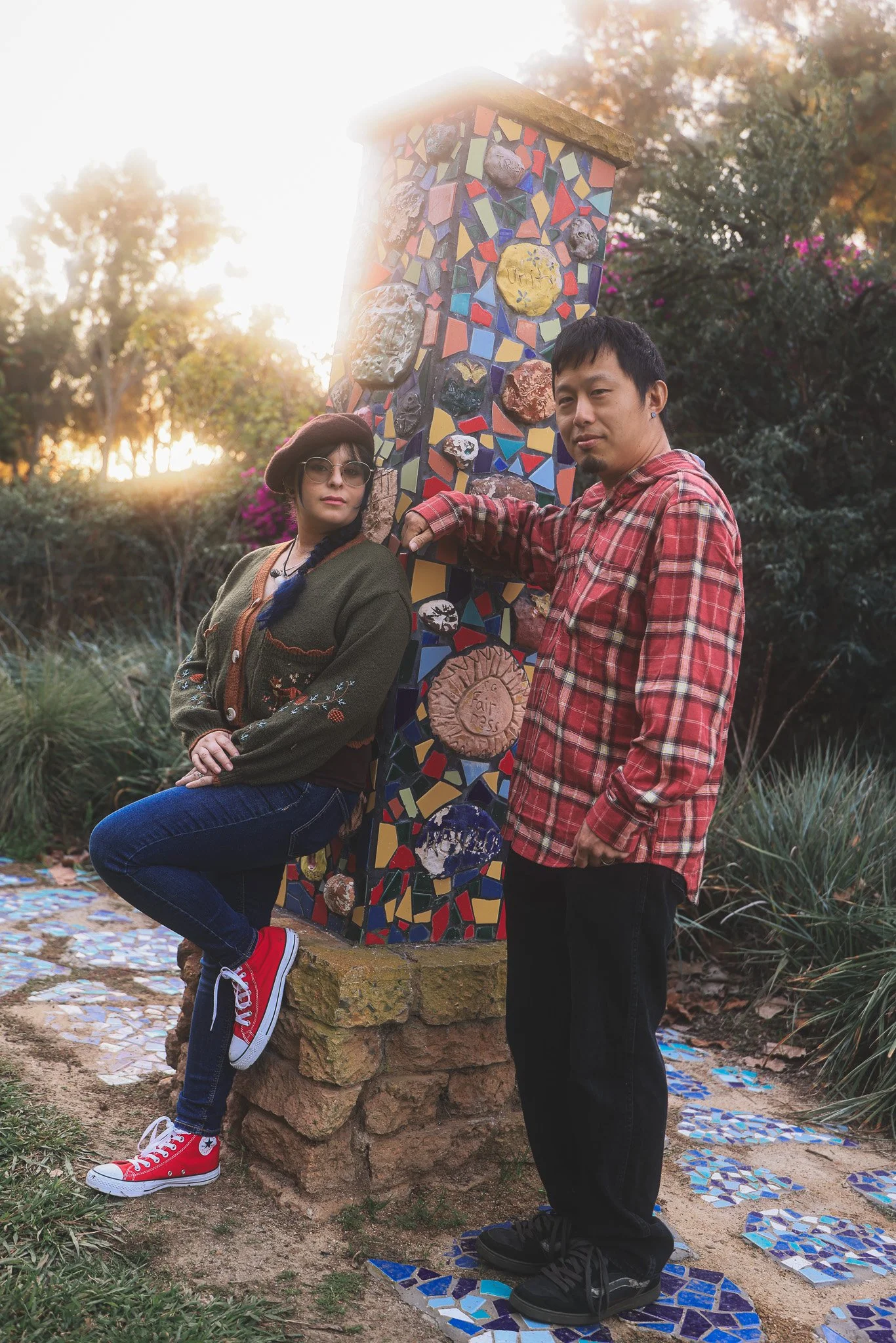 A woman and a man pose next to a colorful mosaic-covered obelisk in a park at sunset.