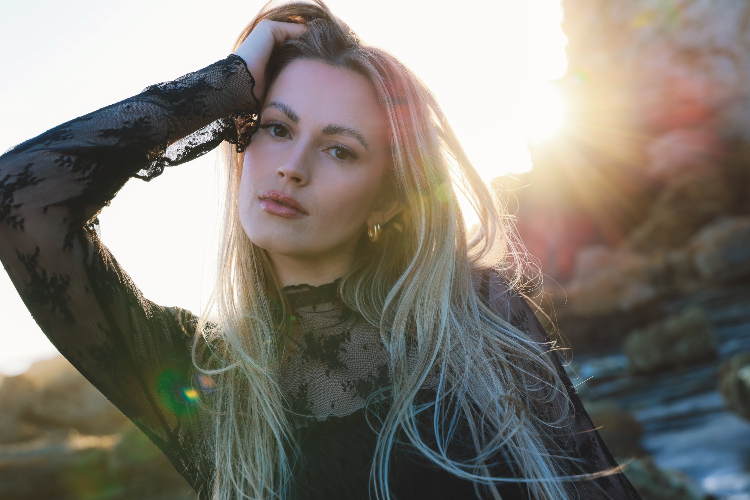 A young woman with long blonde hair, wearing a black lace top, posing outdoors in sunlight with her hand resting on her head.