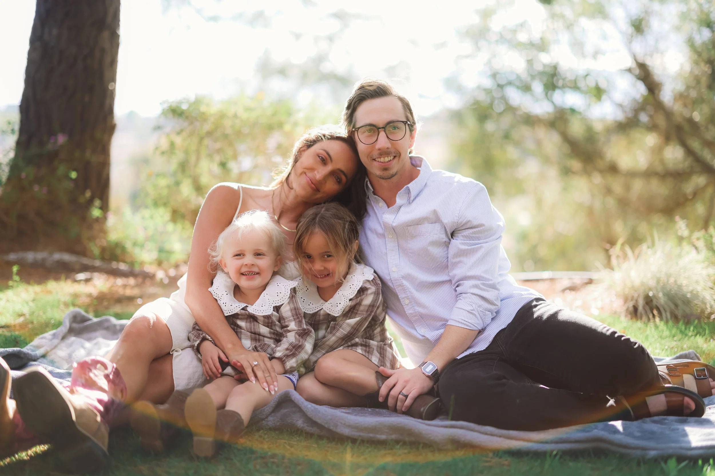 A family of four sitting on a blanket outdoors in a park, smiling for a photo. The parents are behind, with the mother resting her head on the father's shoulder. The two young girls are sitting in front of them, smiling.