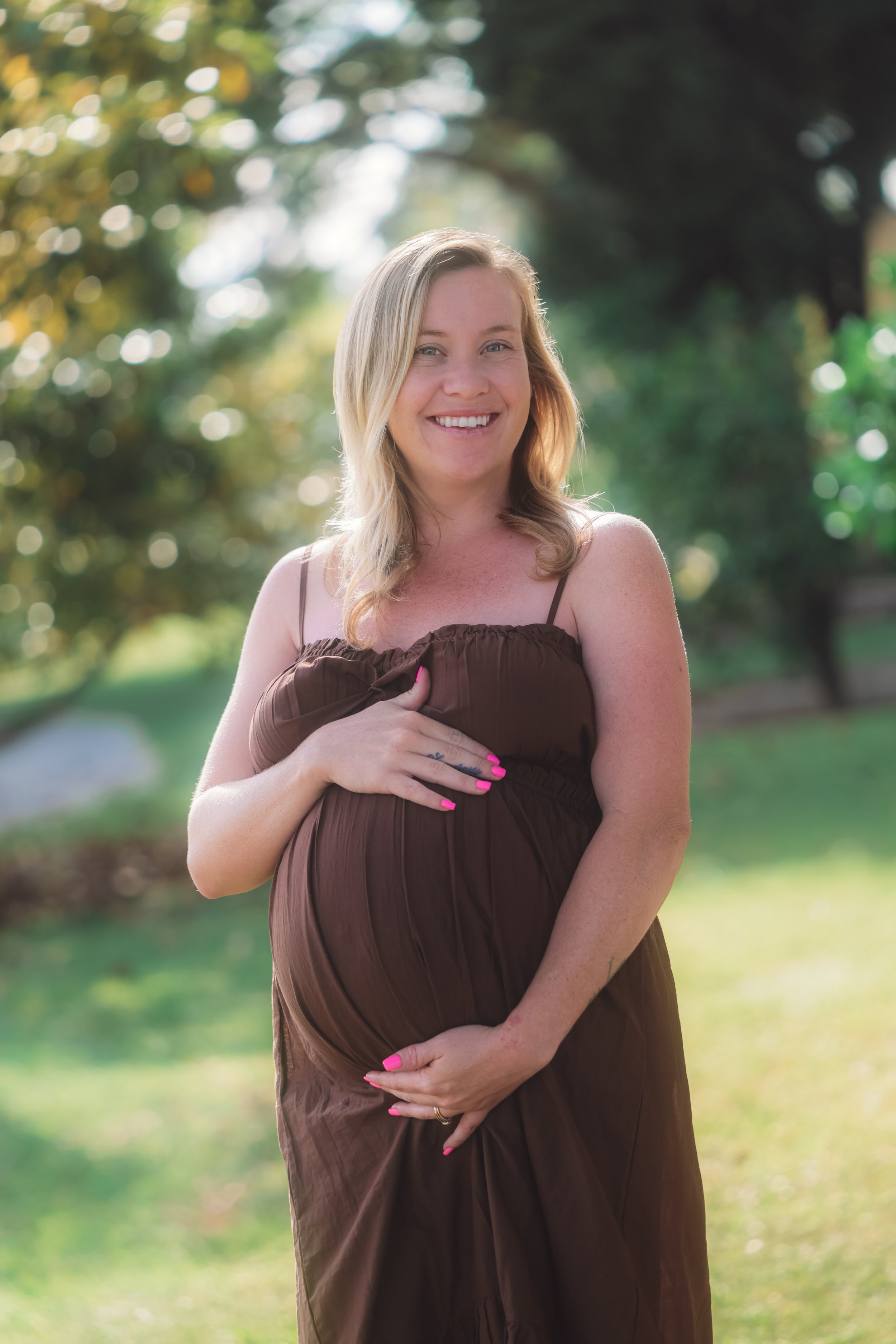 A smiling pregnant woman with blonde hair, wearing a brown dress, standing outdoors in a green park during daylight.