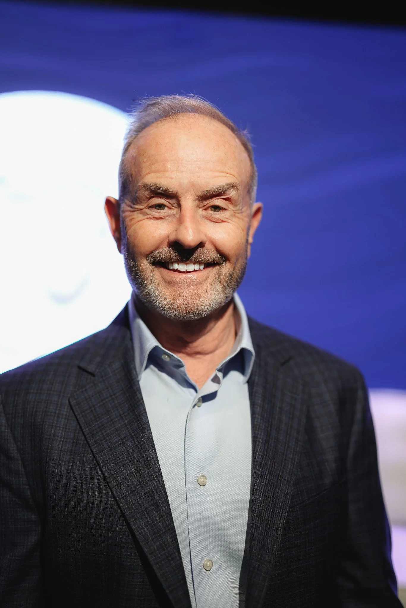 A middle-aged man with a beard and gray hair, smiling while wearing a gray checkered blazer and a light blue collared shirt, standing against a blue background with a white circular light.