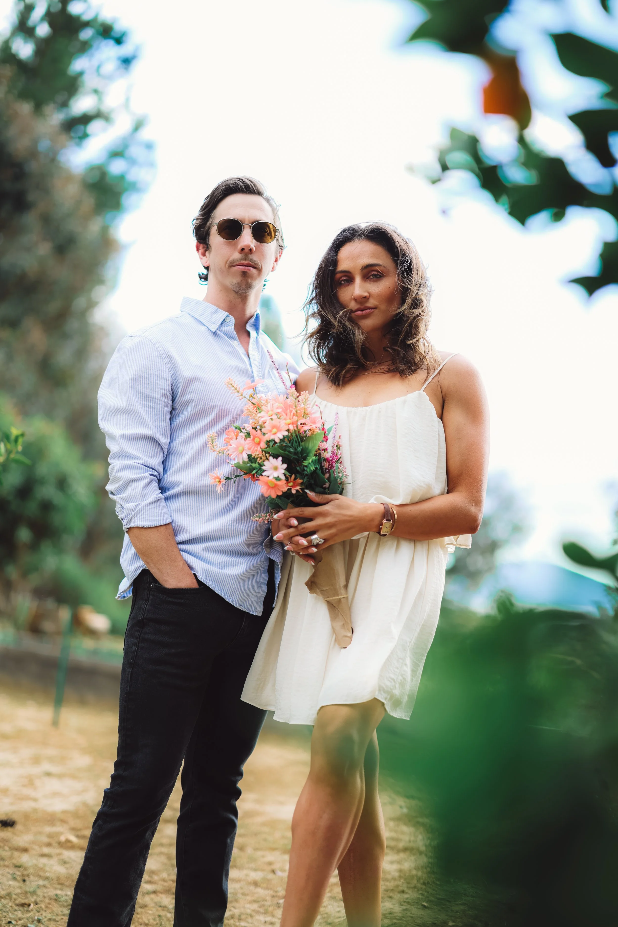 A man and woman standing outdoors, with the woman holding a bouquet of pink flowers. The man is wearing sunglasses, a light blue button-up shirt, and dark jeans. The woman is wearing a white dress with thin straps, has wavy brown hair, and is wearing