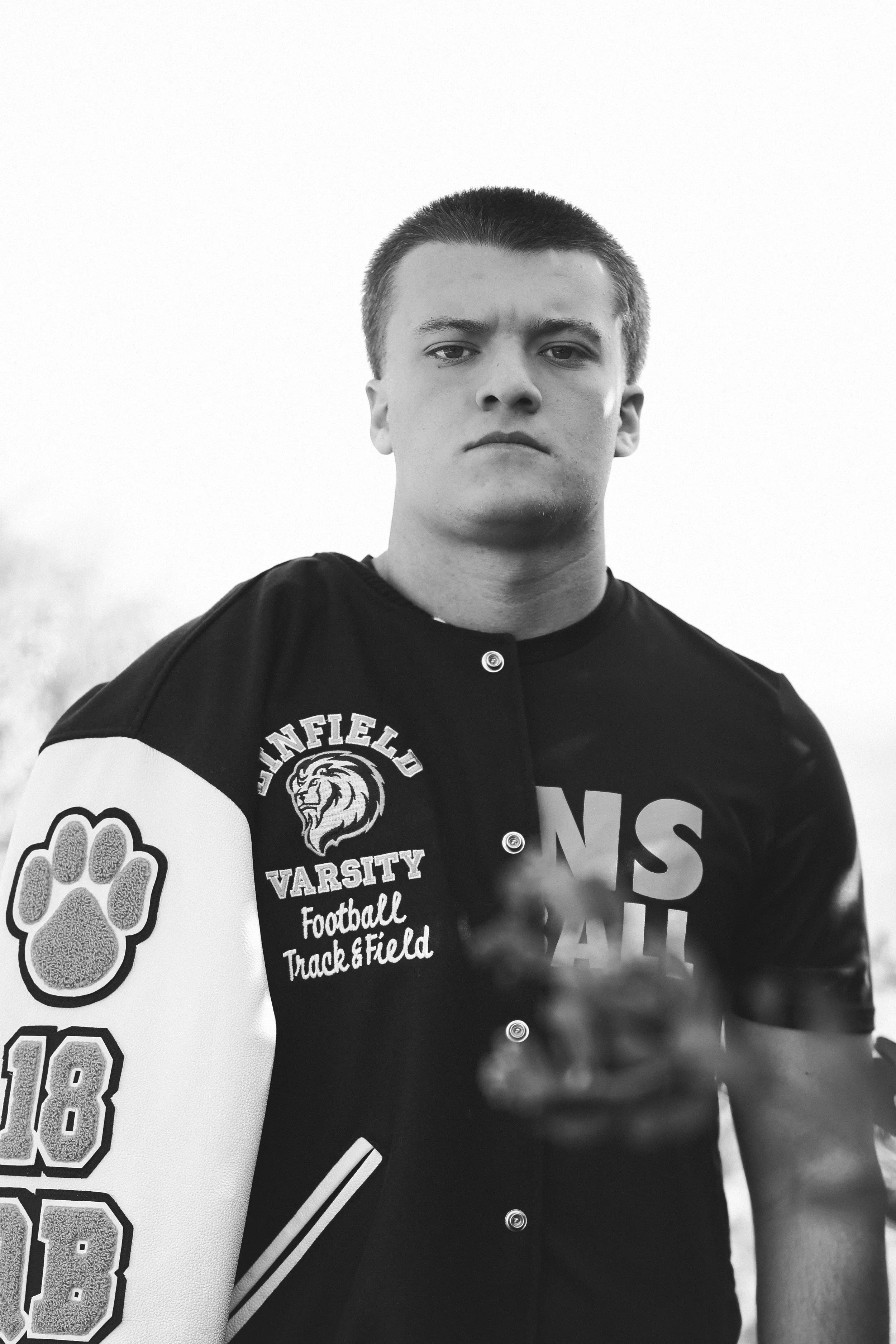 A young man wearing a varsity football jacket with a large paw print and the number 18 on the sleeve, standing outdoors with a serious expression.