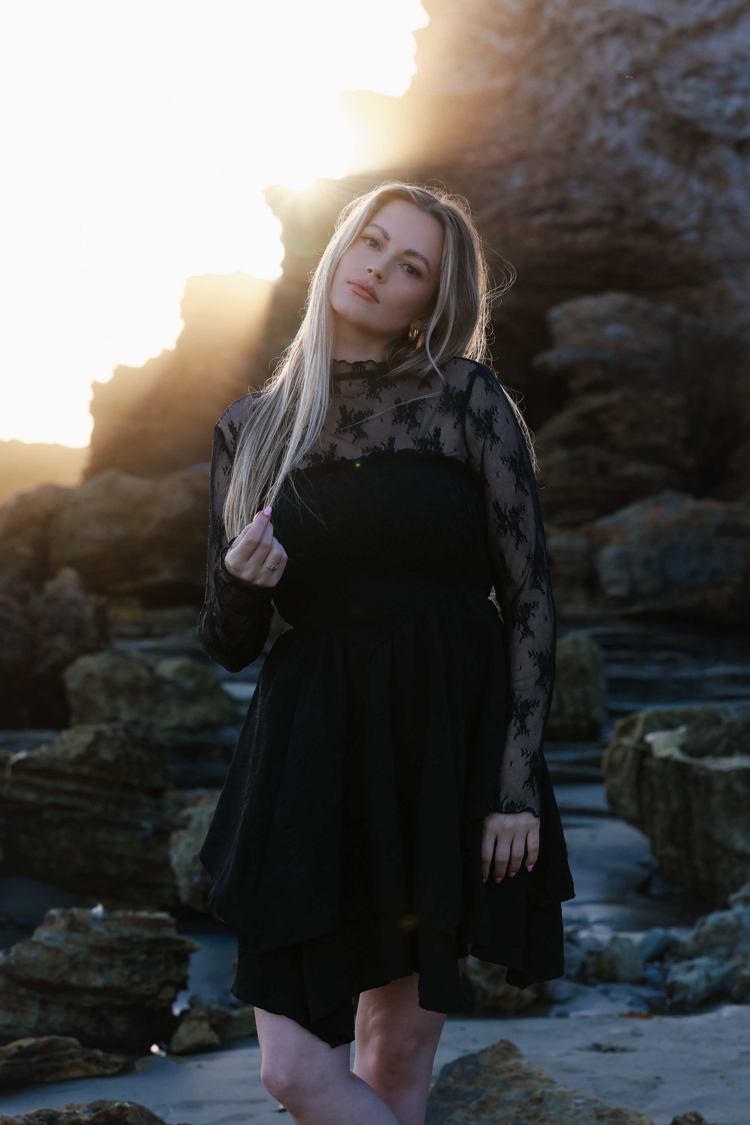 A young woman in a black lace dress standing on rocks at the beach during sunset.