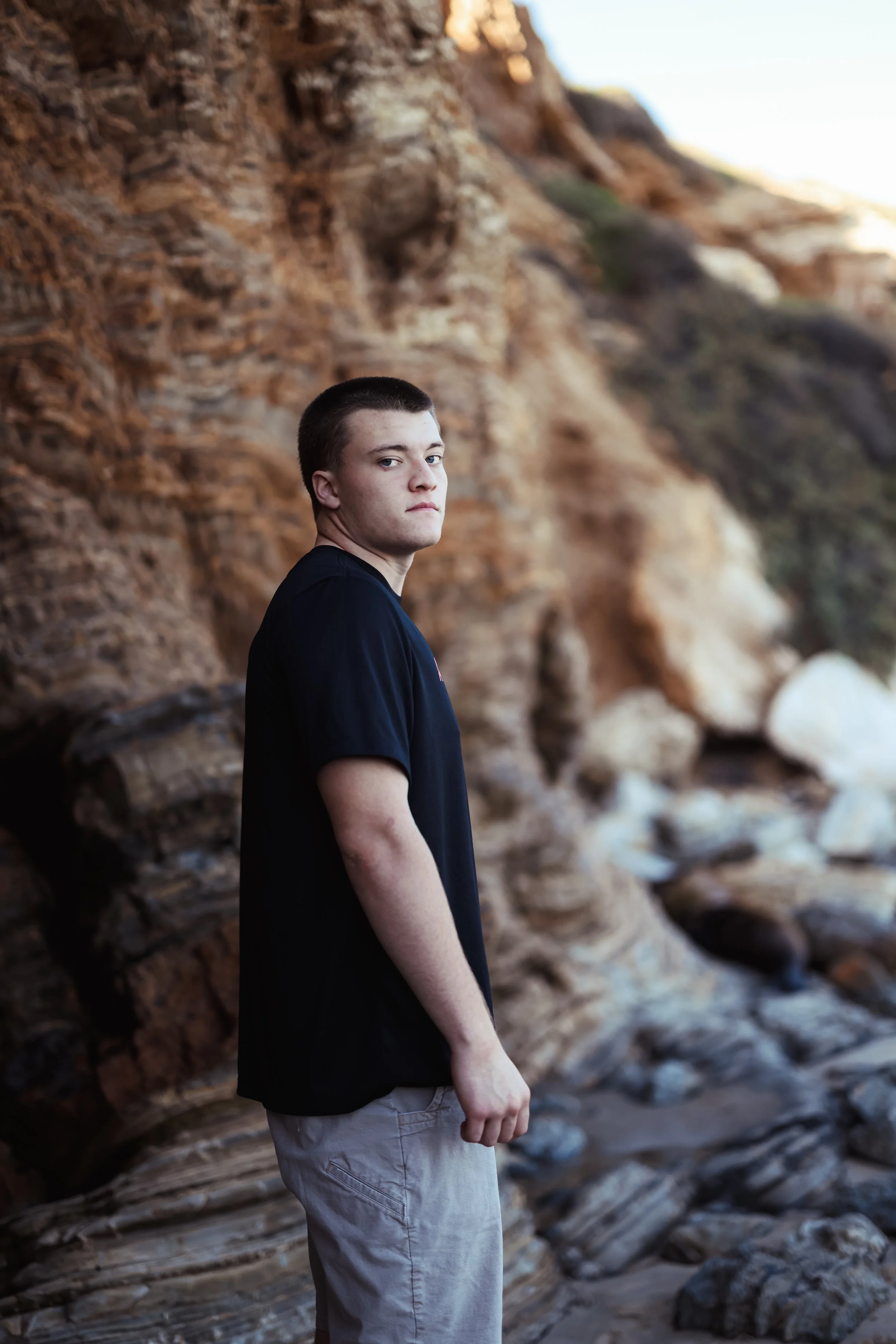 A young man with short dark hair standing near rocky cliffs at the beach, looking at the camera with a serious expression.