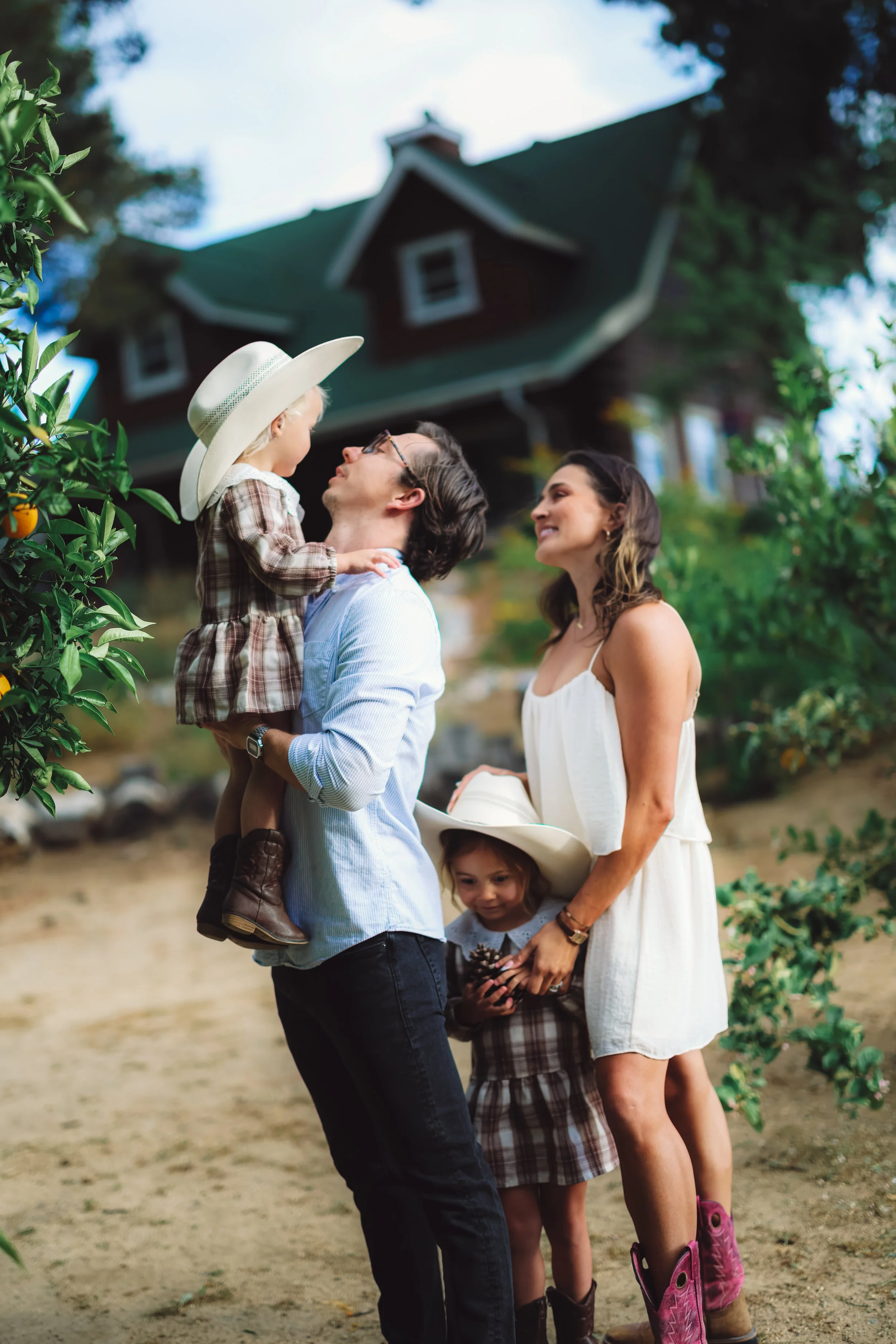 A family of four outdoors, with a man holding a young girl wearing a large wide-brimmed hat and boots, with a woman in a white dress and a young girl in a plaid dress holding pinecones, all smiling and looking happily at each other. A house and trees