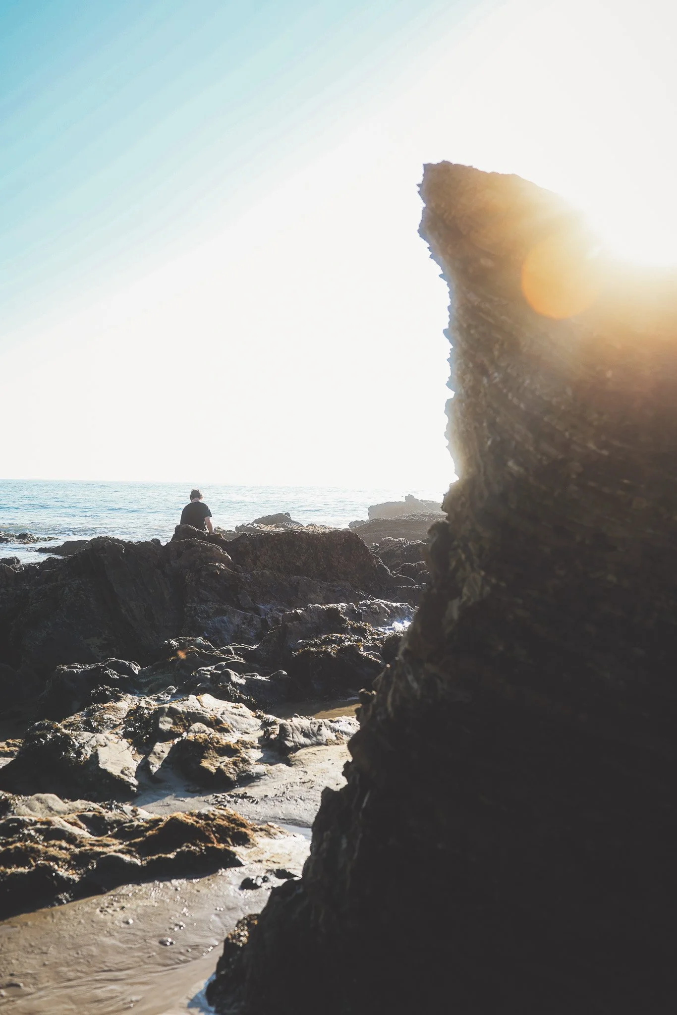 A person sitting on rocks at the beach, facing the ocean, with large rock formations around and sunlight shining from behind, creating a lens flare.