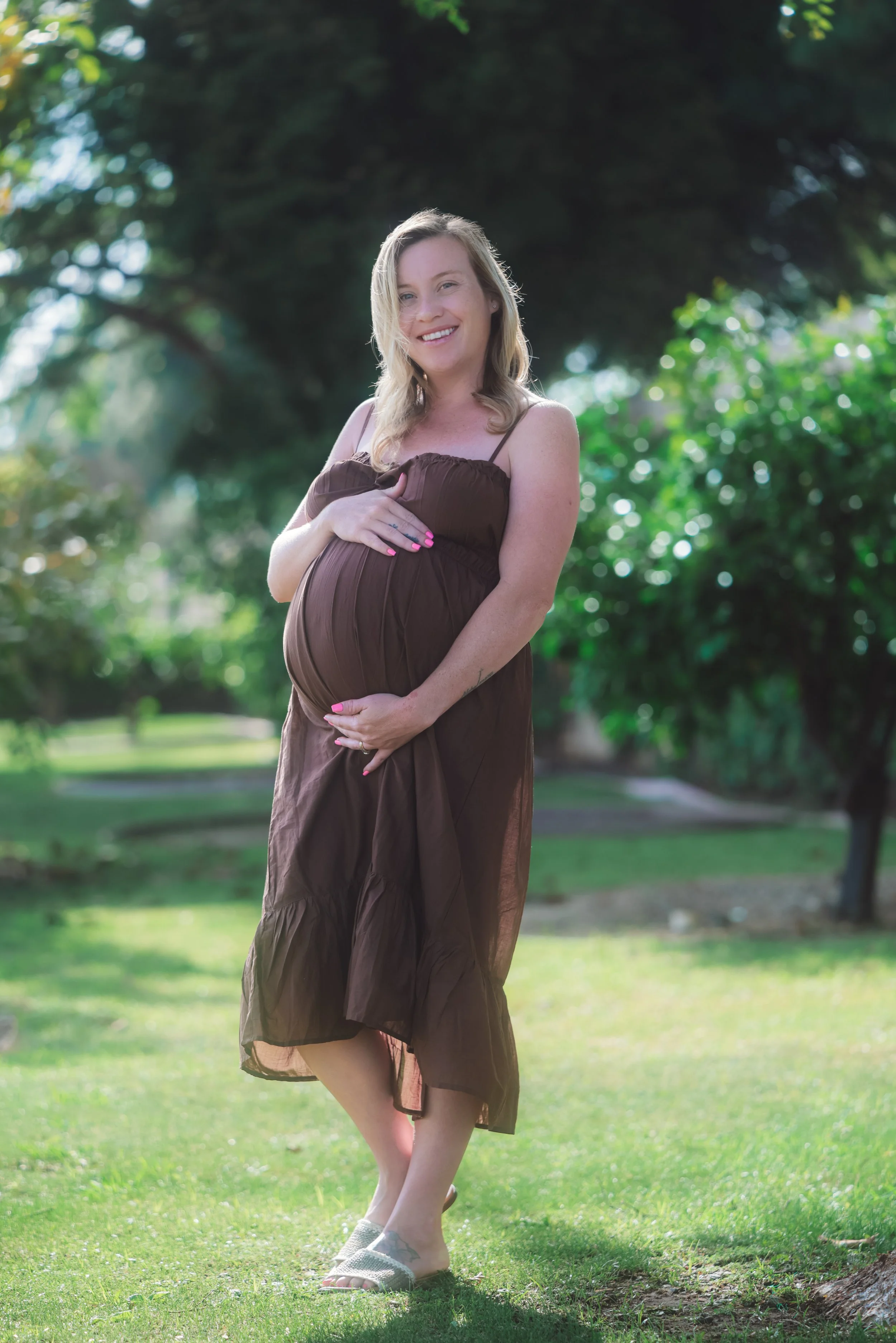 A pregnant woman in a brown dress smiling outdoors on a sunny day surrounded by green trees.
