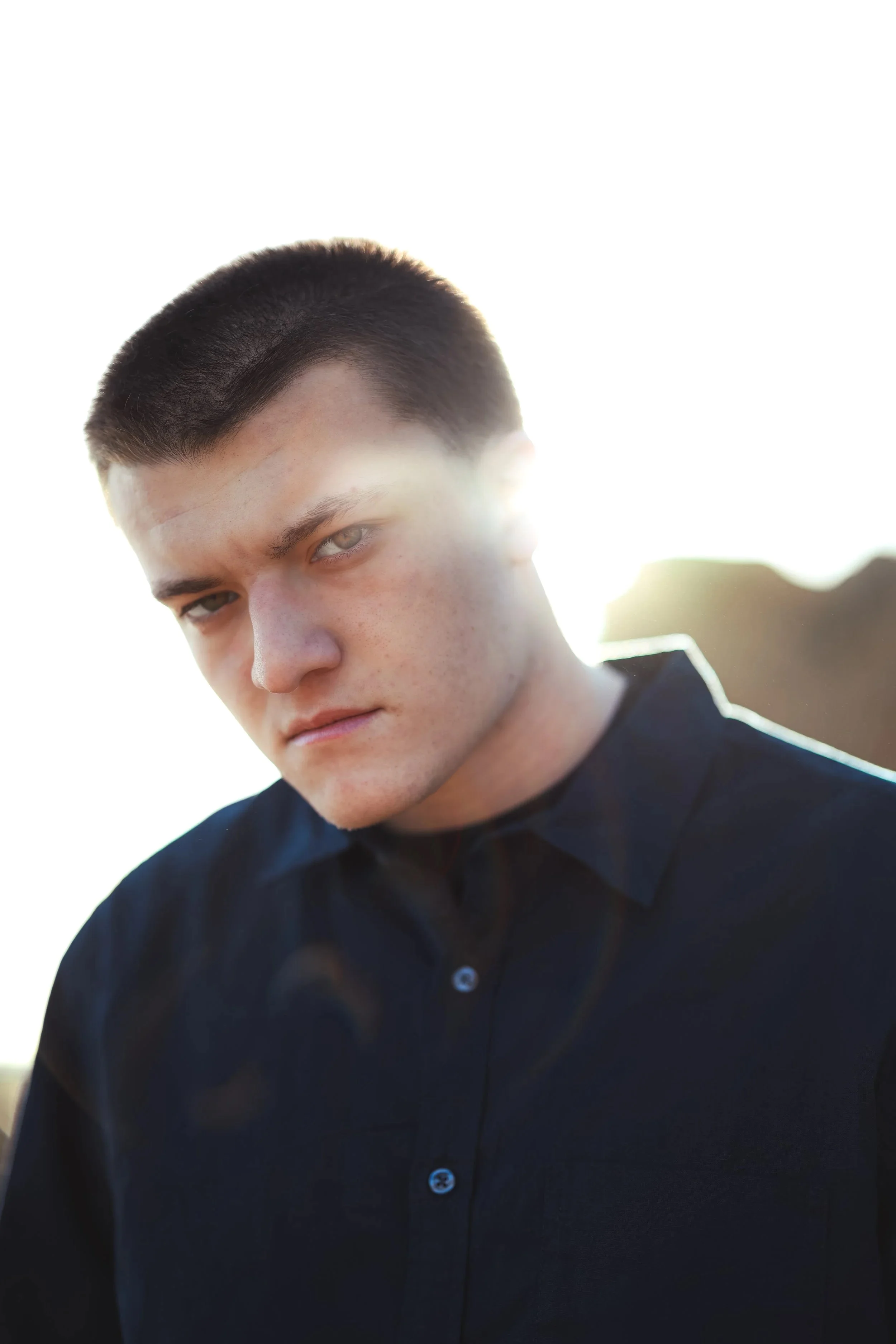 A young man with short dark hair and blue eyes looking serious, wearing a dark shirt, with bright sunlight and lens flare in the background.