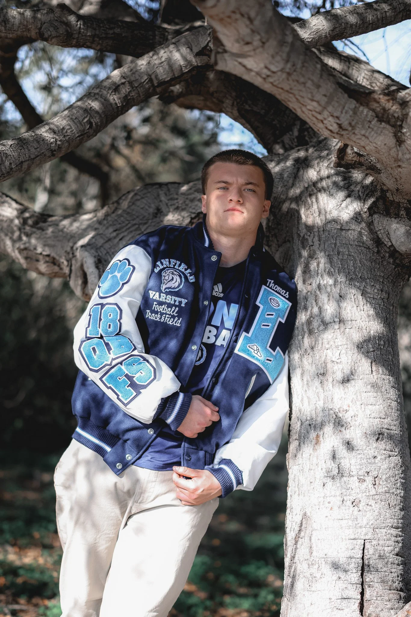 A young man leaning against a large tree with a thick trunk and sprawling branches, outdoors during the day.