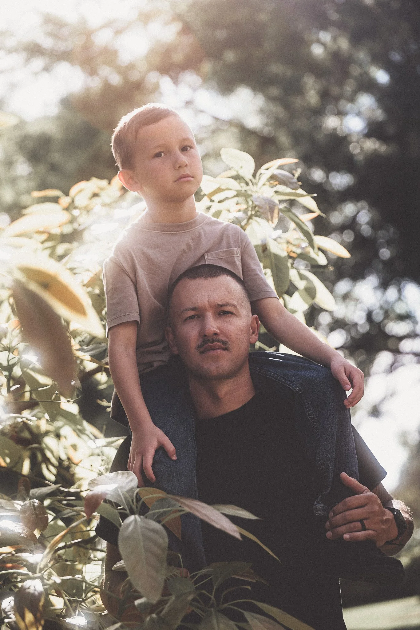 A man carrying a boy on his shoulders outside among green leaves, with sunlight filtering through the trees.