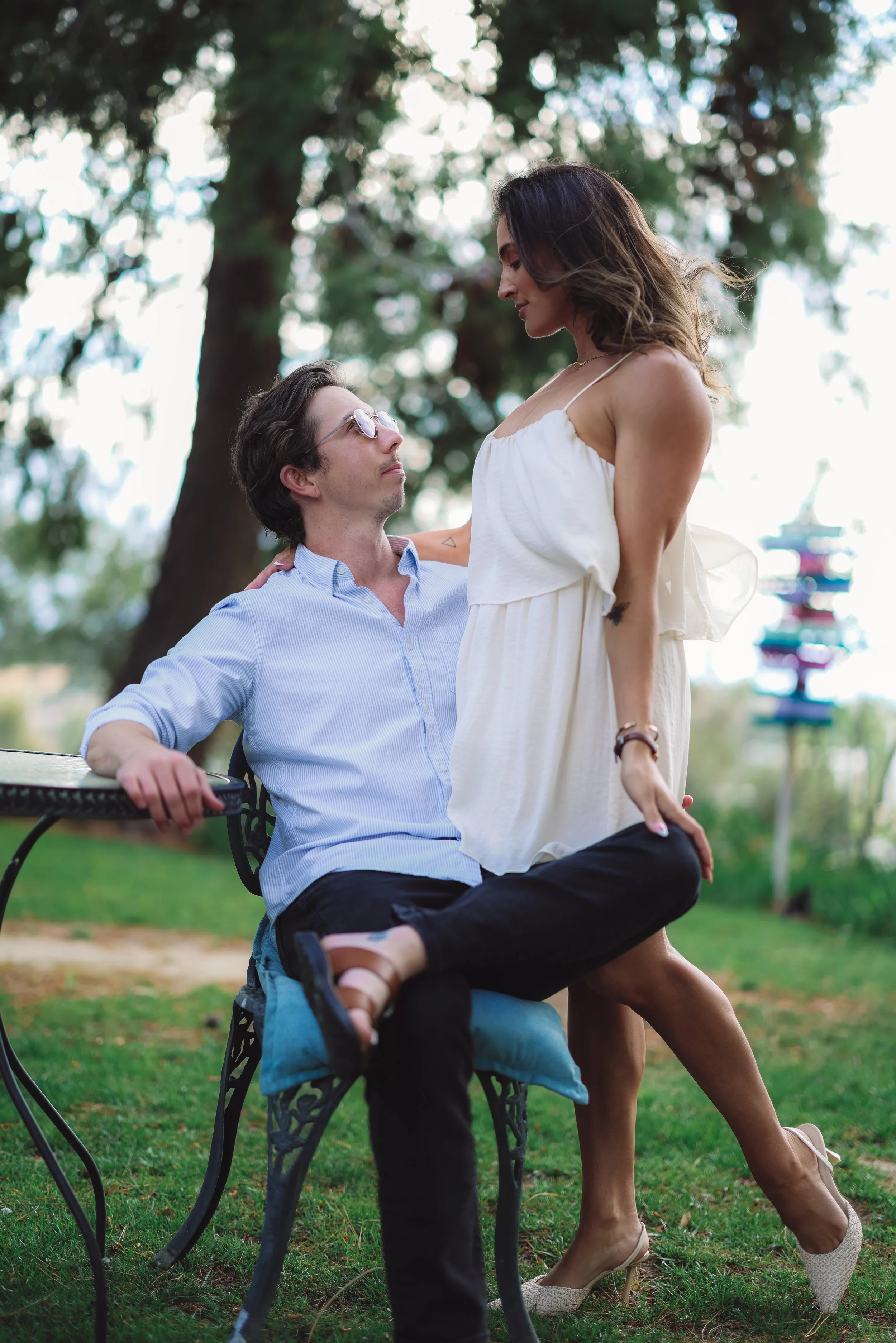 A man and woman having a romantic moment outdoors in a park with trees, grass, and a blurred background of a colorful sculpture. The man is sitting on a bench, looking up at the woman who is standing close to him.