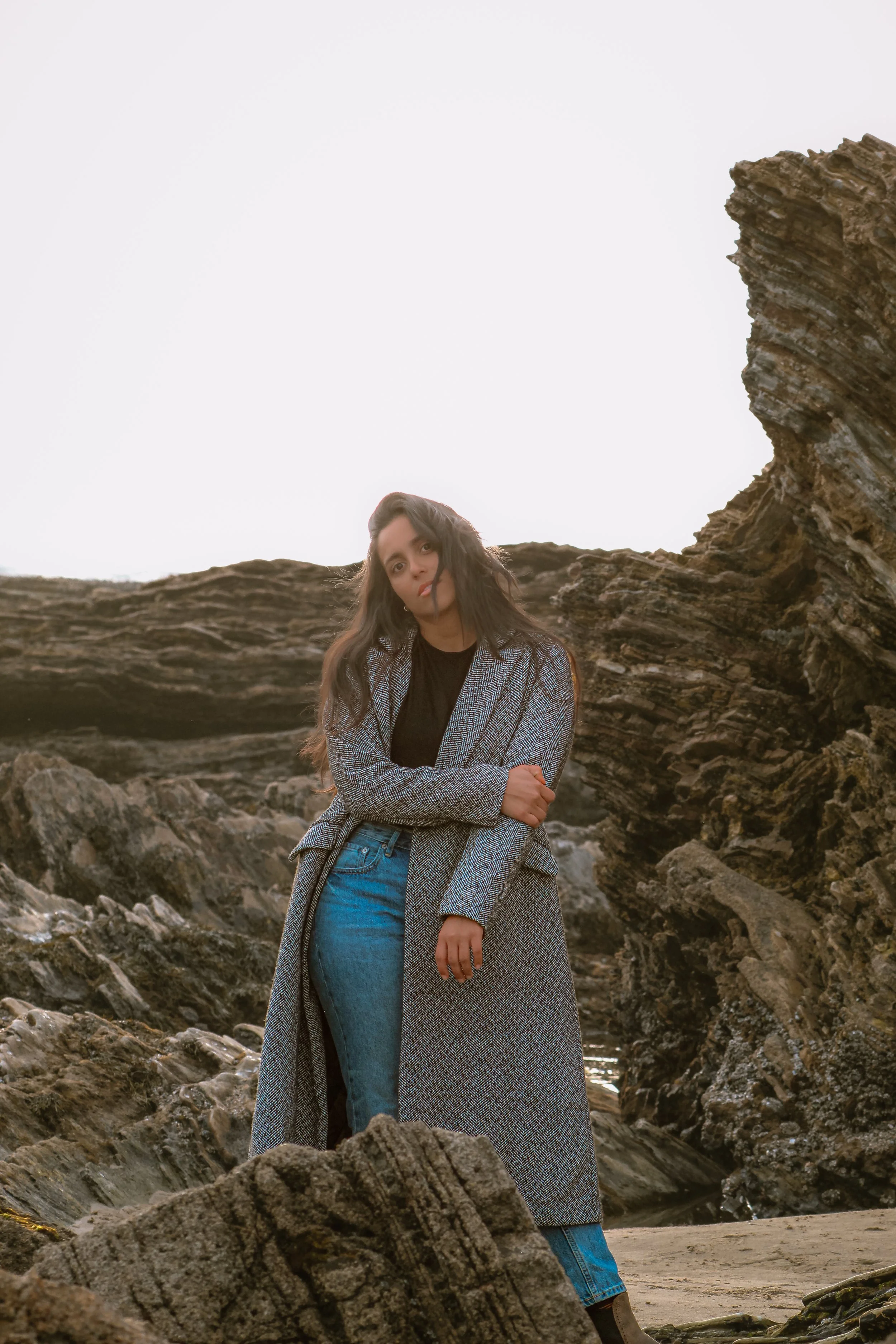 A woman with long dark hair wearing a gray coat, black top, and blue jeans standing among rocks on a beach with a large textured rock formation behind her and a bright overcast sky.