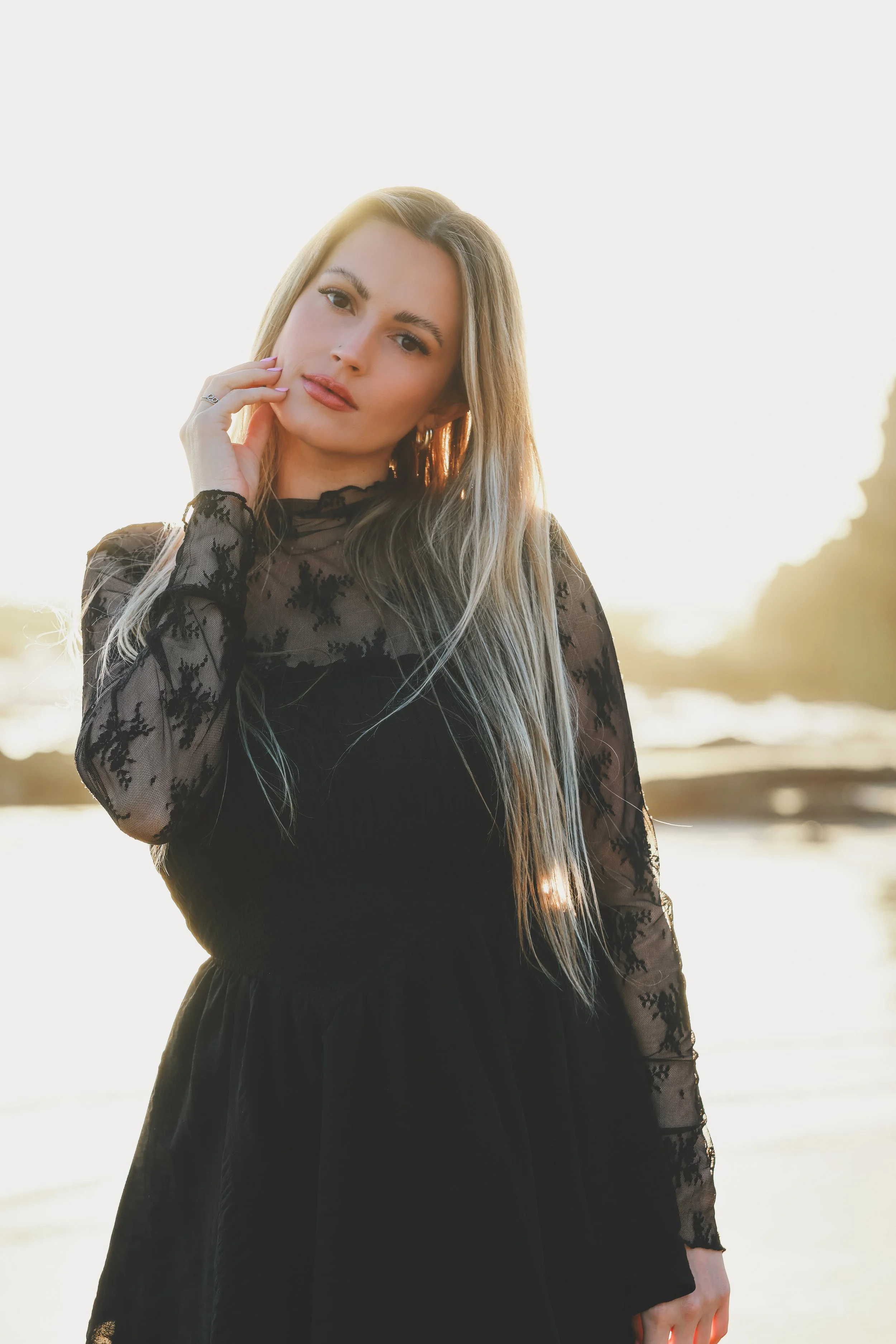 A young woman with long blonde hair wearing a black dress with lace sleeves, standing outdoors during sunset near a body of water.