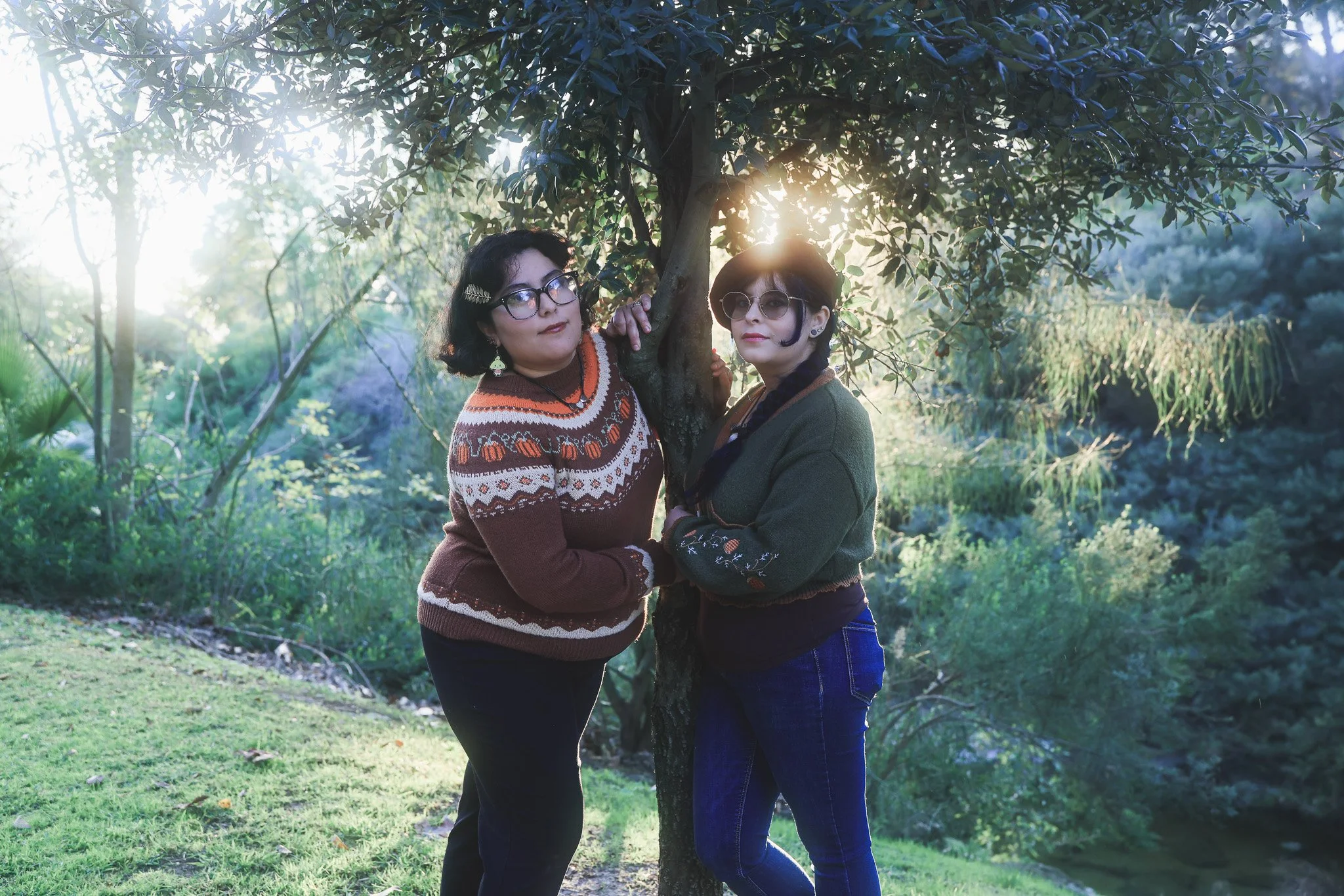Two women standing under a tree in a park with sunlight shining through the branches, one with dark curly hair and glasses, the other with short dark hair and sunglasses, both wearing sweaters.