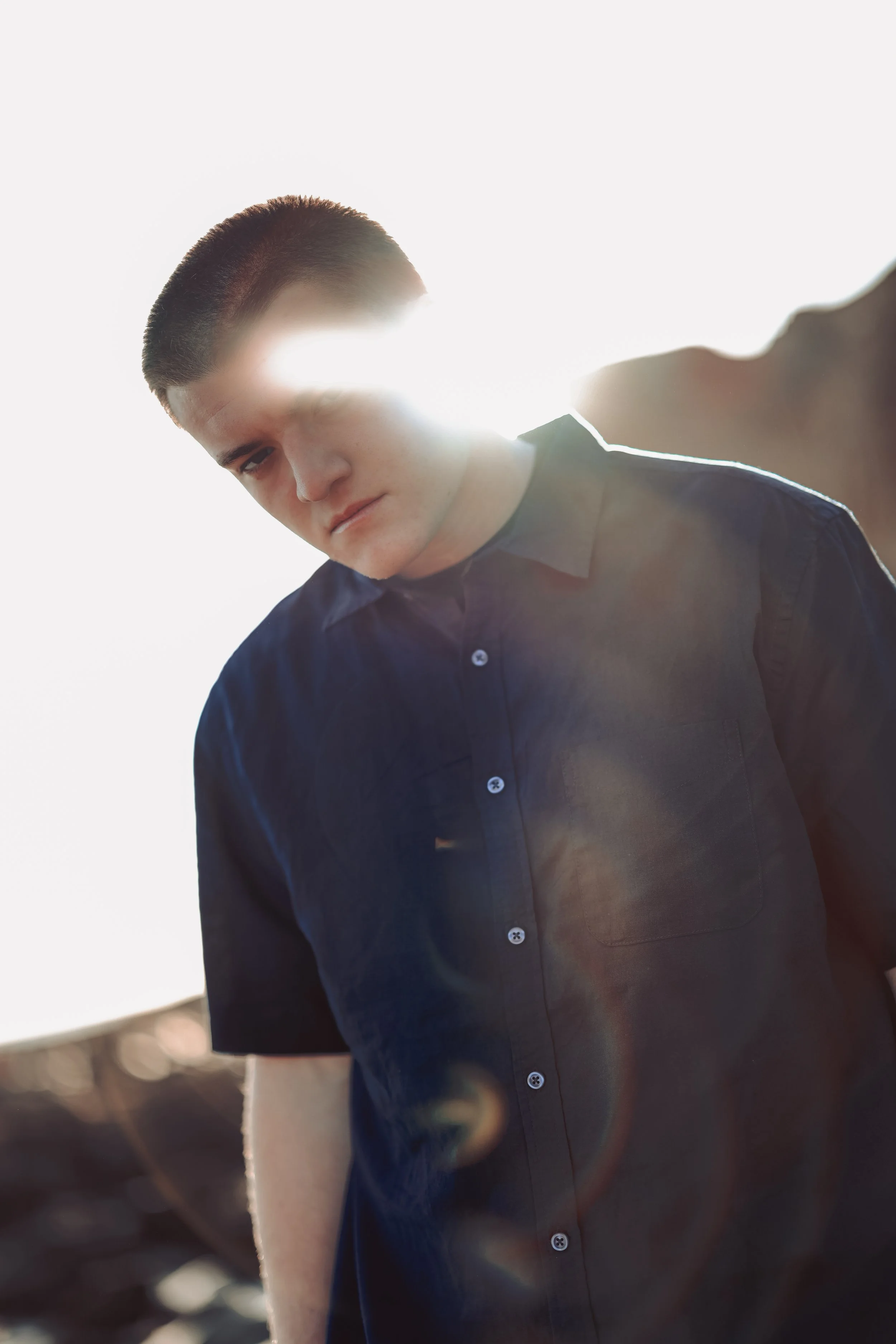 Young man with short dark hair wearing a black shirt, looking down, with sunlight behind him creating a lens flare effect.