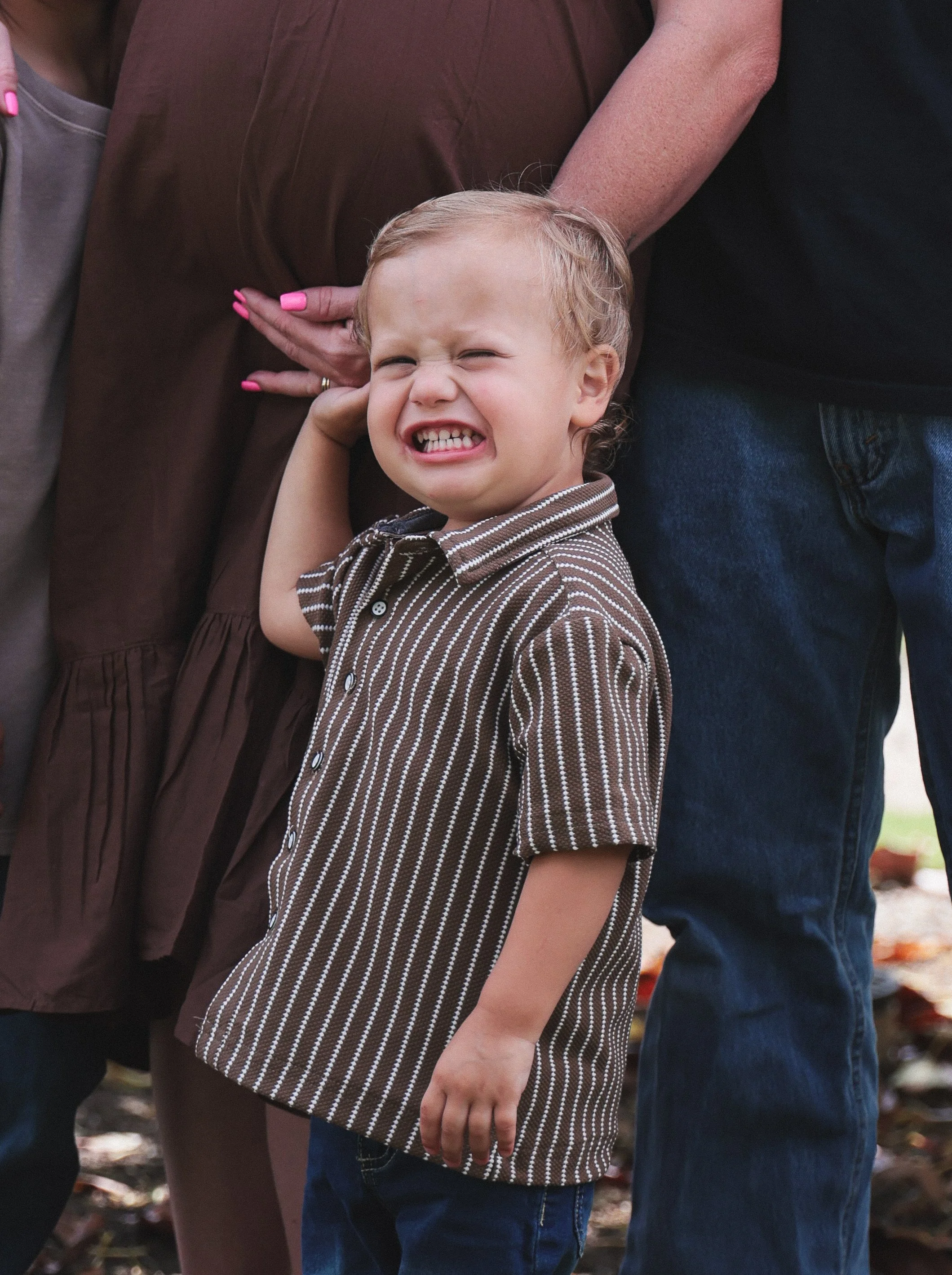 A young boy with blond hair and wearing a brown striped shirt is grimacing and squinting, with one hand resting on his chin, standing outdoors with three adults partially visible around him.
