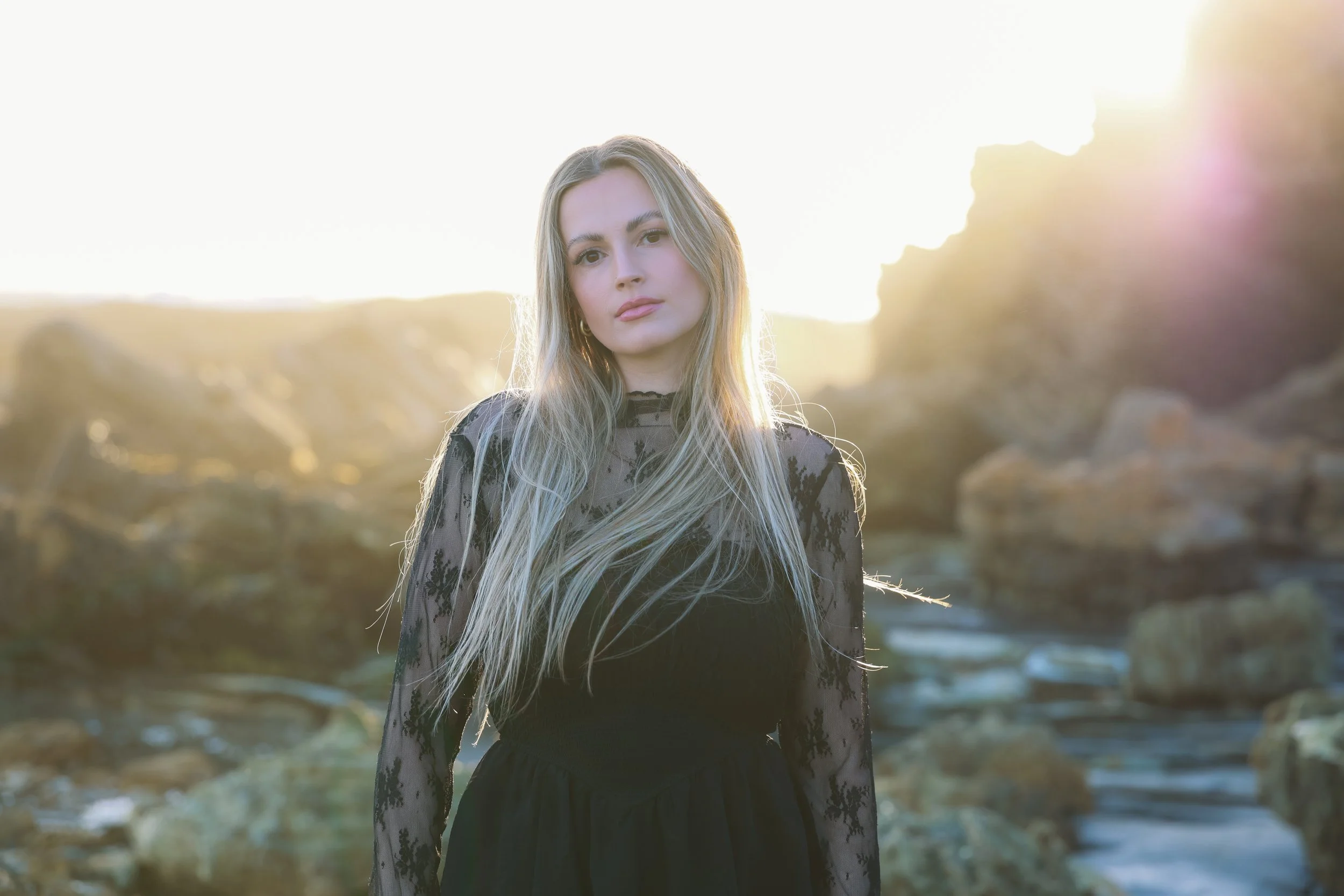 A young woman with long blonde hair wearing a black lace dress standing outdoors near rocks with sunlight in the background.