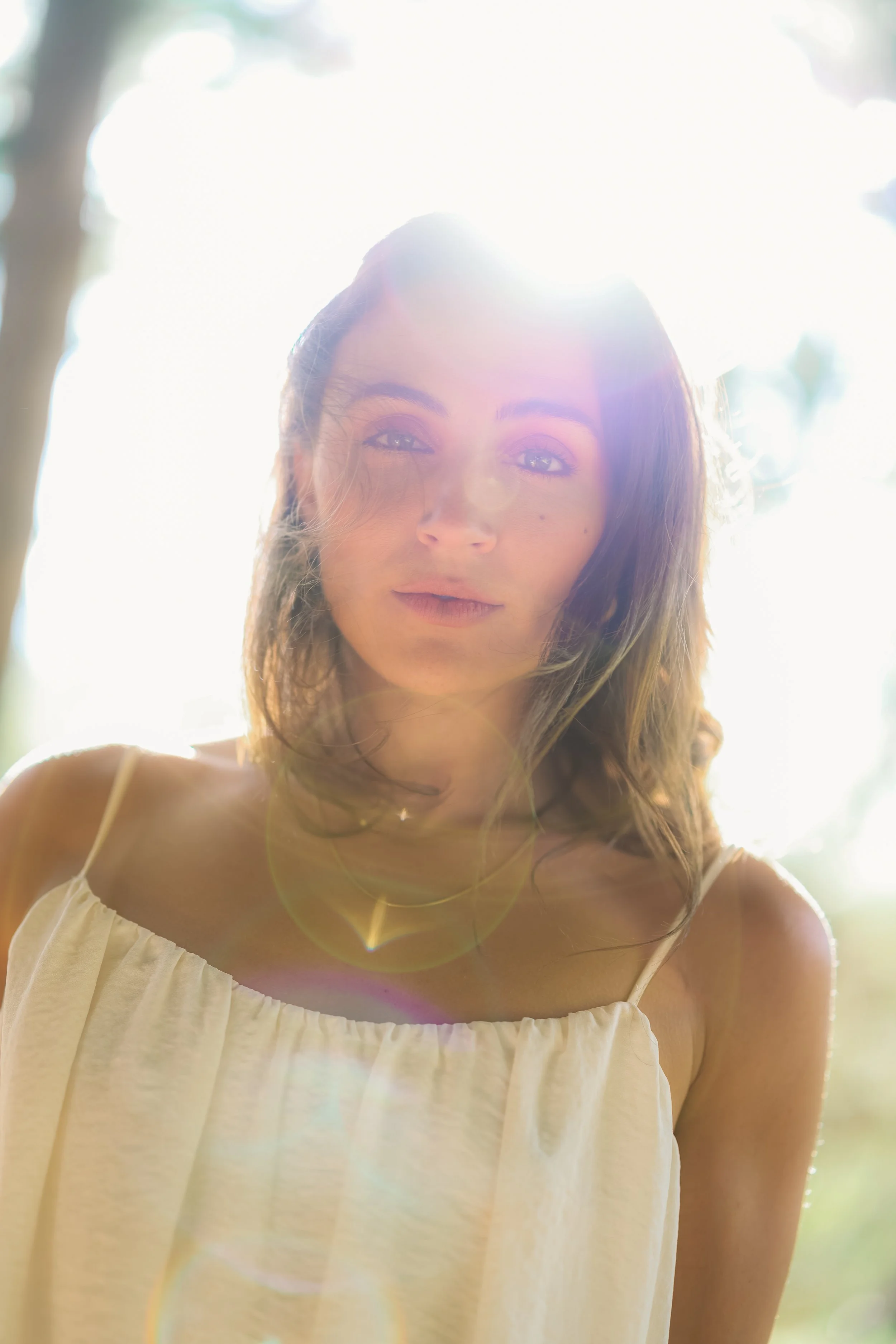 Young woman with shoulder-length brown hair and light-colored eyes, wearing a white sleeveless top, standing outdoors with sunlight shining behind her.