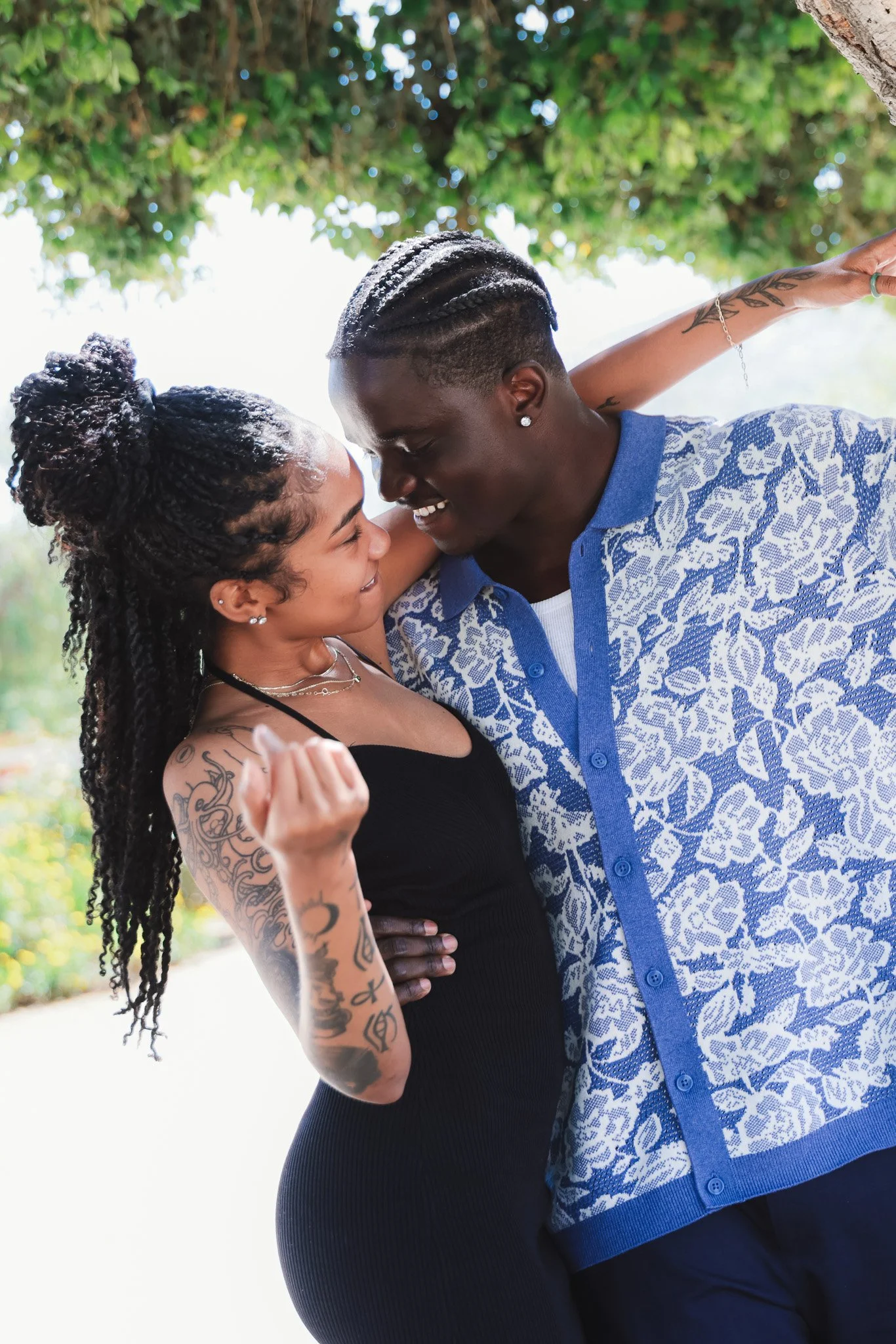 A couple is dancing outdoors, smiling and close together, under a leafy green tree.