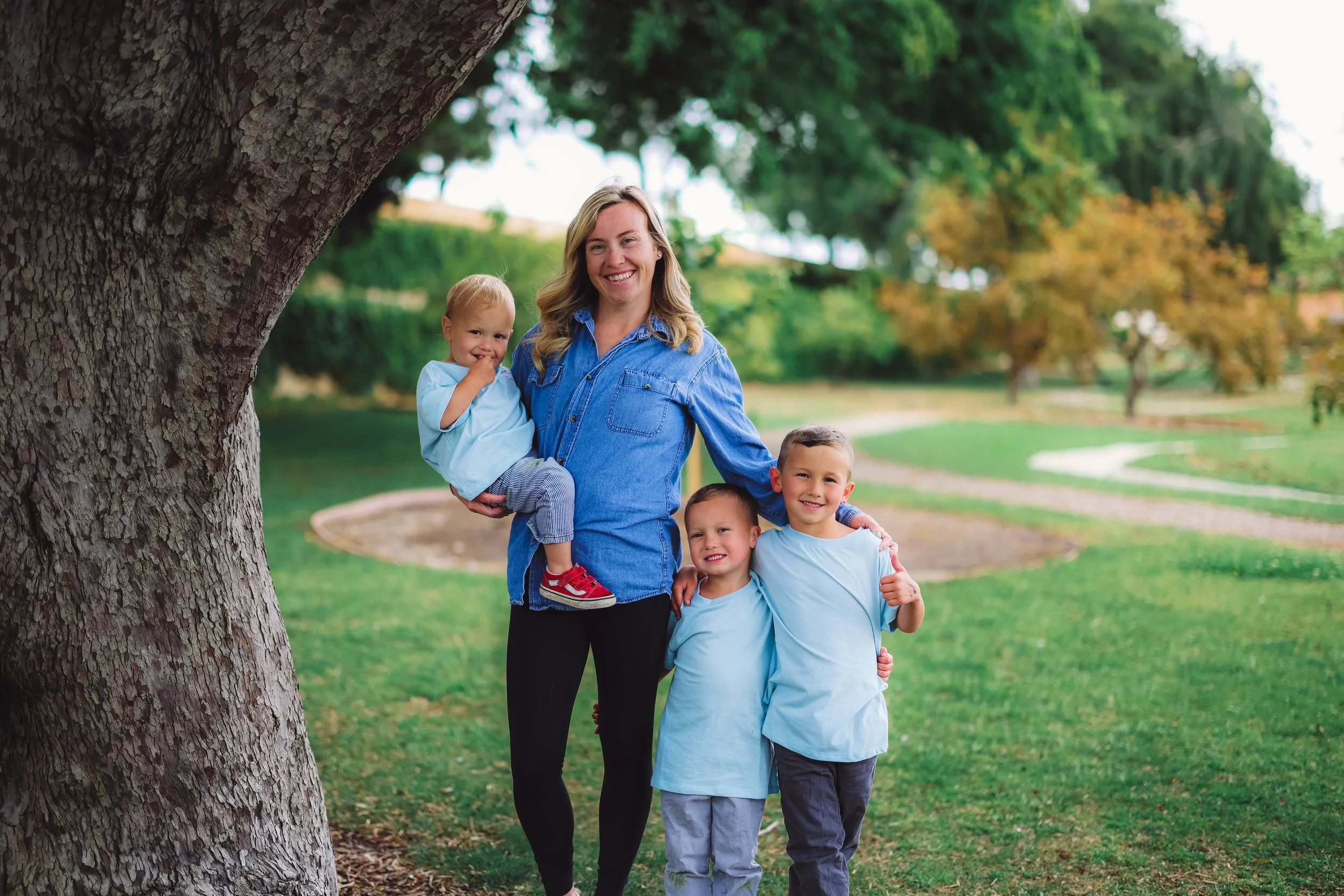 A smiling woman with blonde hair standing under a large tree, holding a young girl, and hugging two young boys, in a park with green grass and trees in the background.