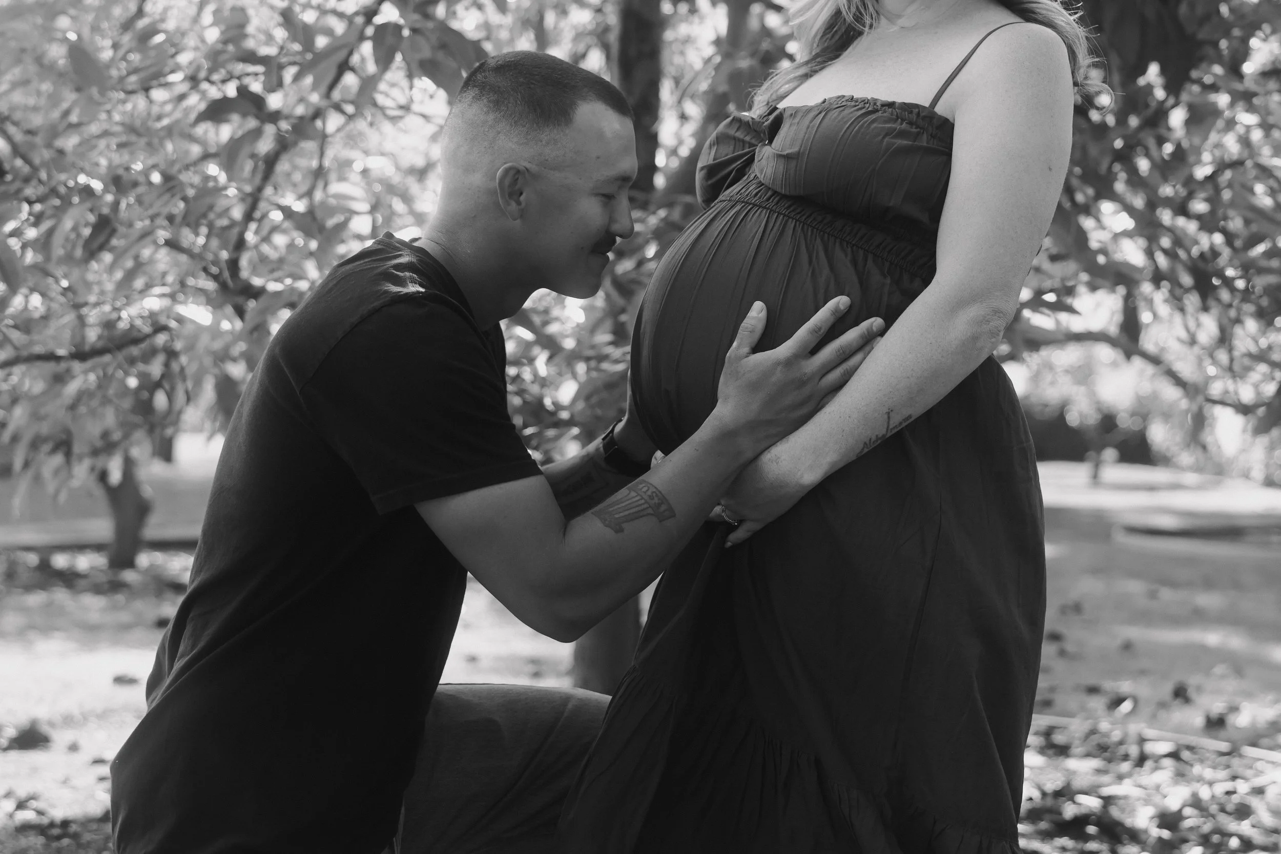 A man kneeling and holding the pregnant belly of a woman, who is standing outdoors with trees in the background, in black and white.