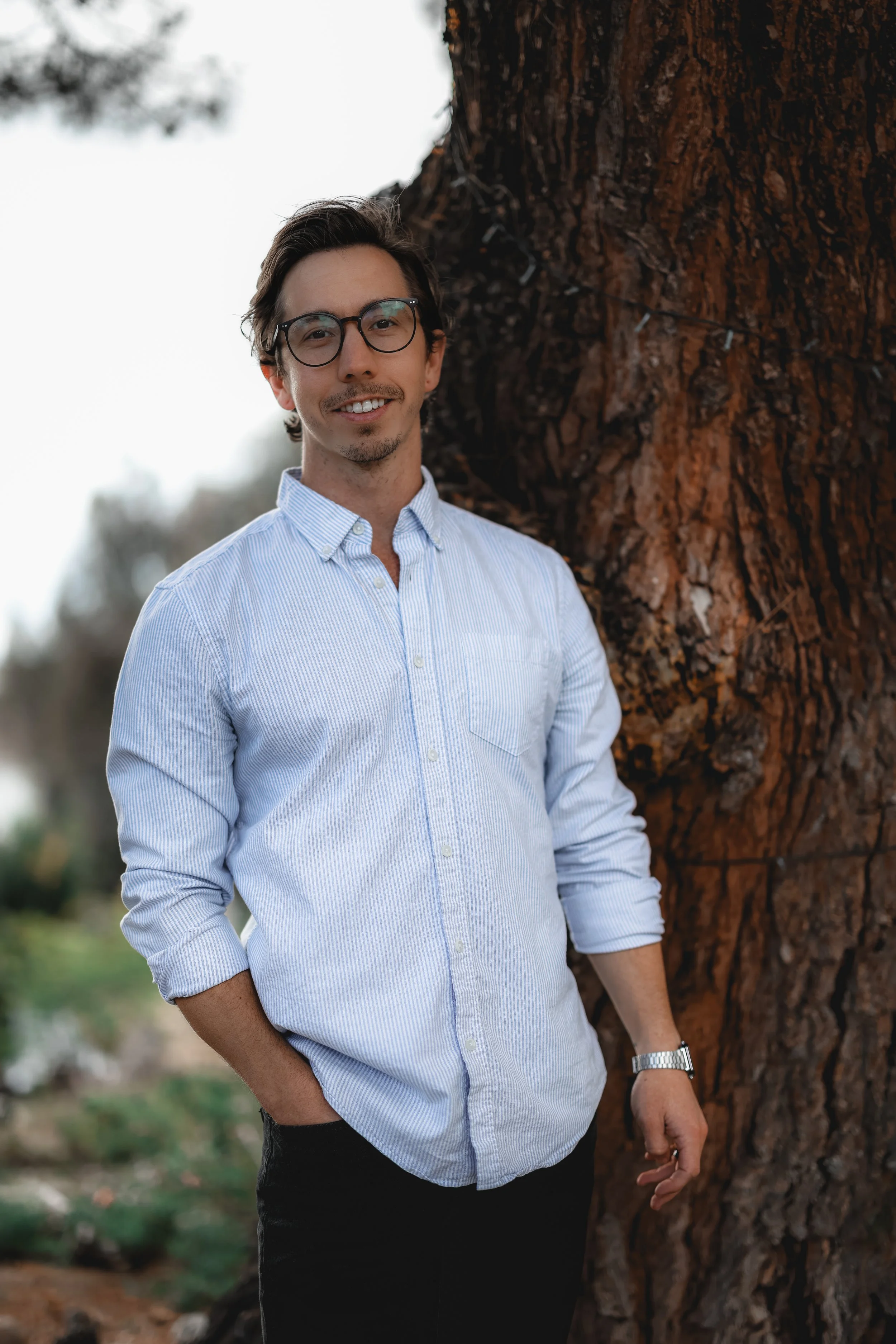 Young man with glasses wearing a light blue button-up shirt, standing outdoors next to a large tree.