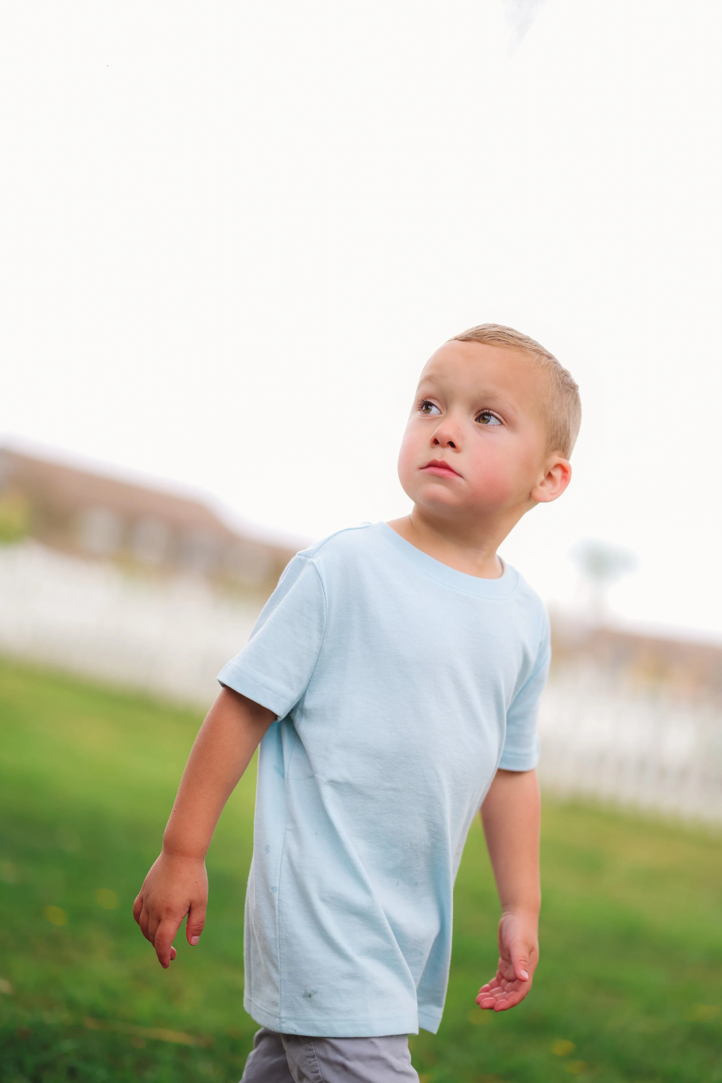 A young boy with short blond hair, wearing a light blue t-shirt, standing outdoors on grass with a blurred background of houses and a white fence, looking to his left with a serious expression.