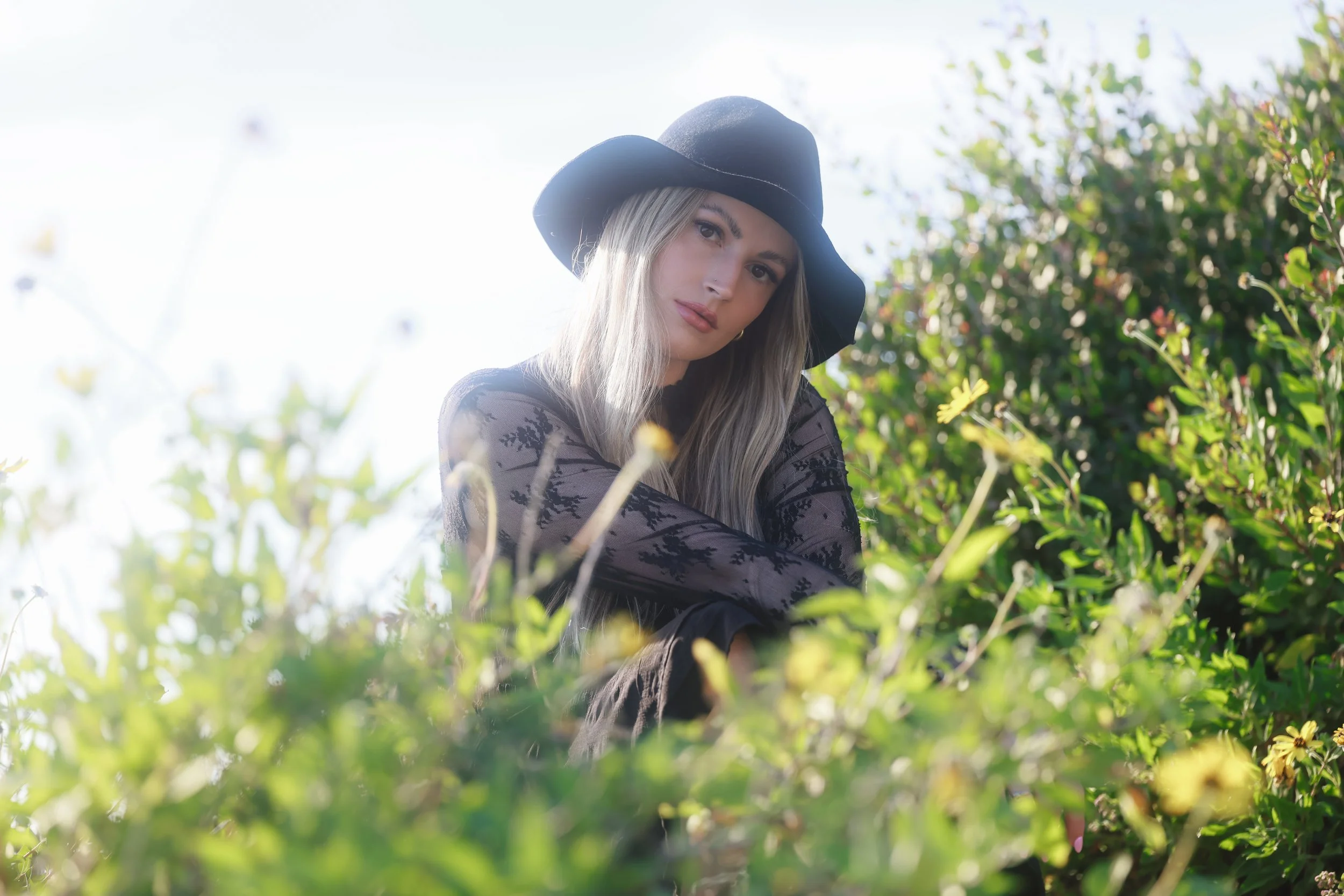 A woman with long blonde hair wearing a black hat and a black lace top sitting among green bushes with yellow flowers, looking at the camera.
