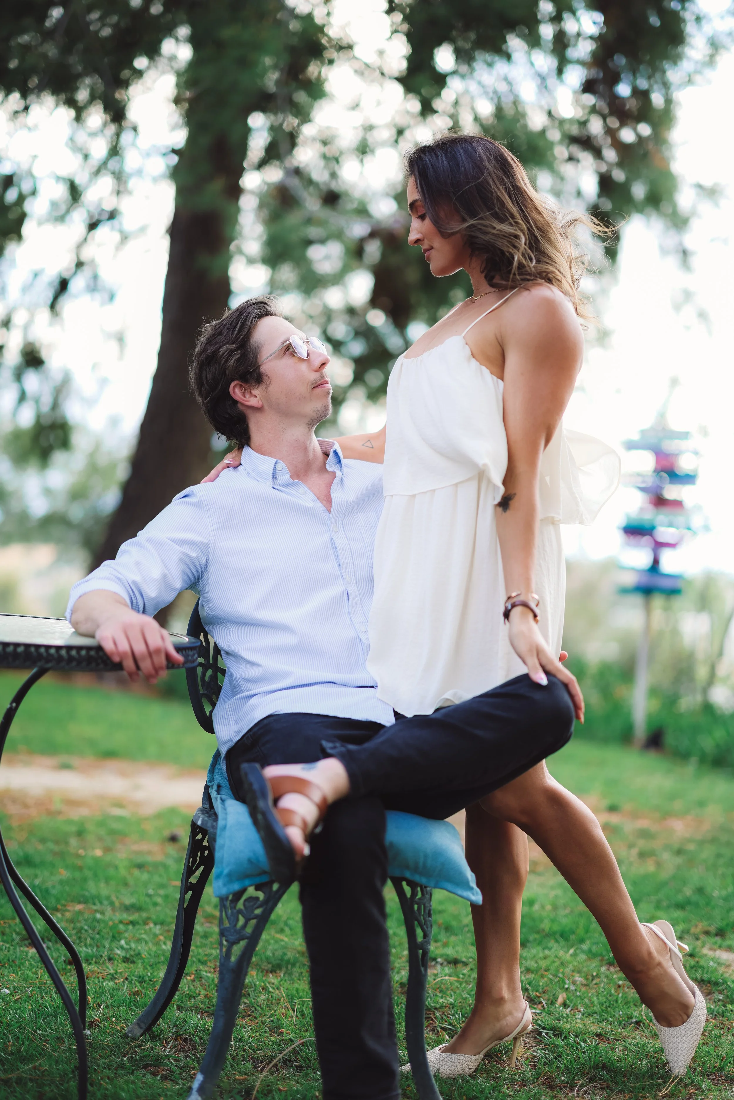 A man sitting on a park bench with sunglasses looking at a woman standing in front of him with her eyes closed. The woman is wearing a white dress and beige heels, while the man is in a light blue shirt. They are outdoors with trees and colorful play