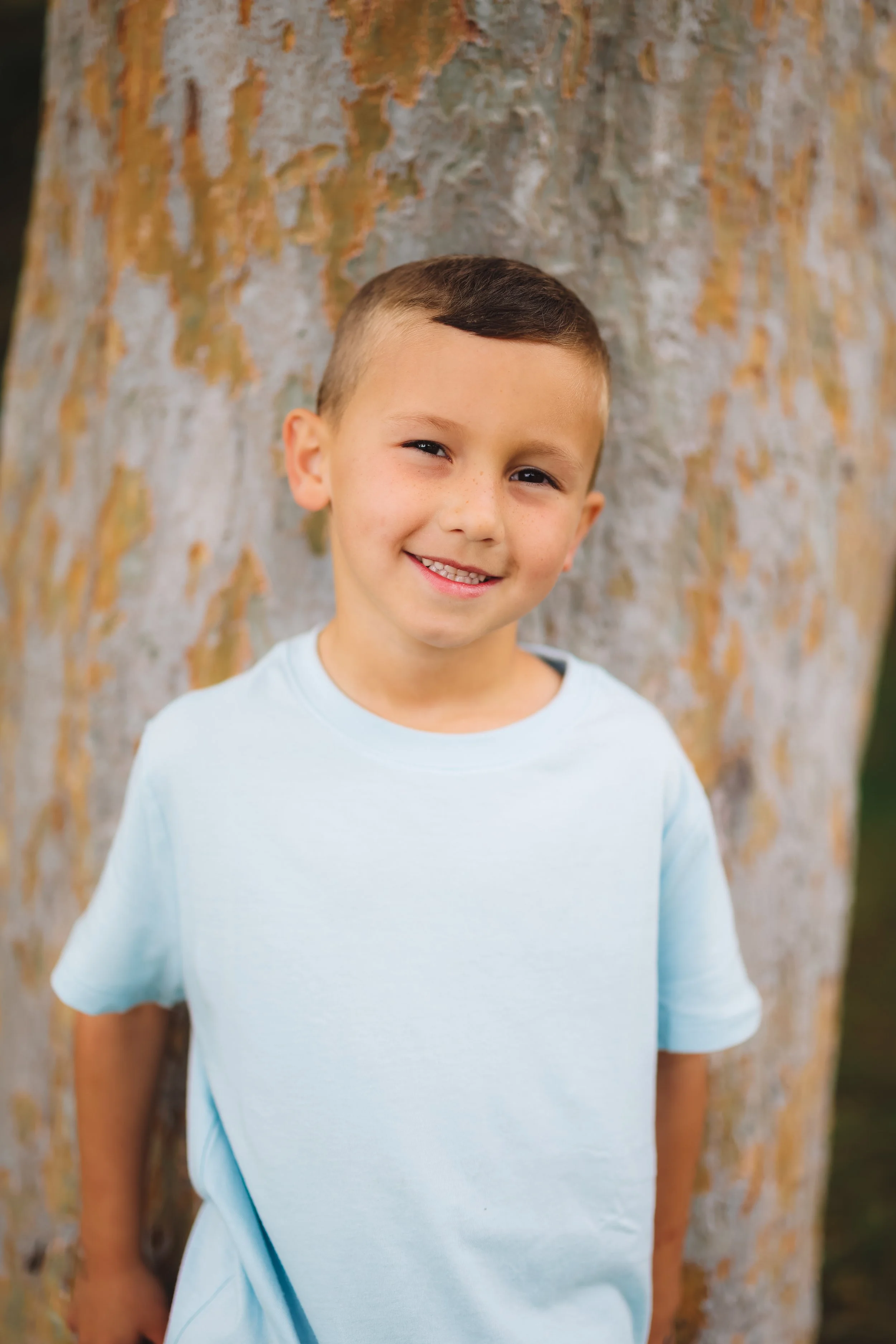 Young boy with short brown hair smiling, standing outdoors in front of a tree trunk.