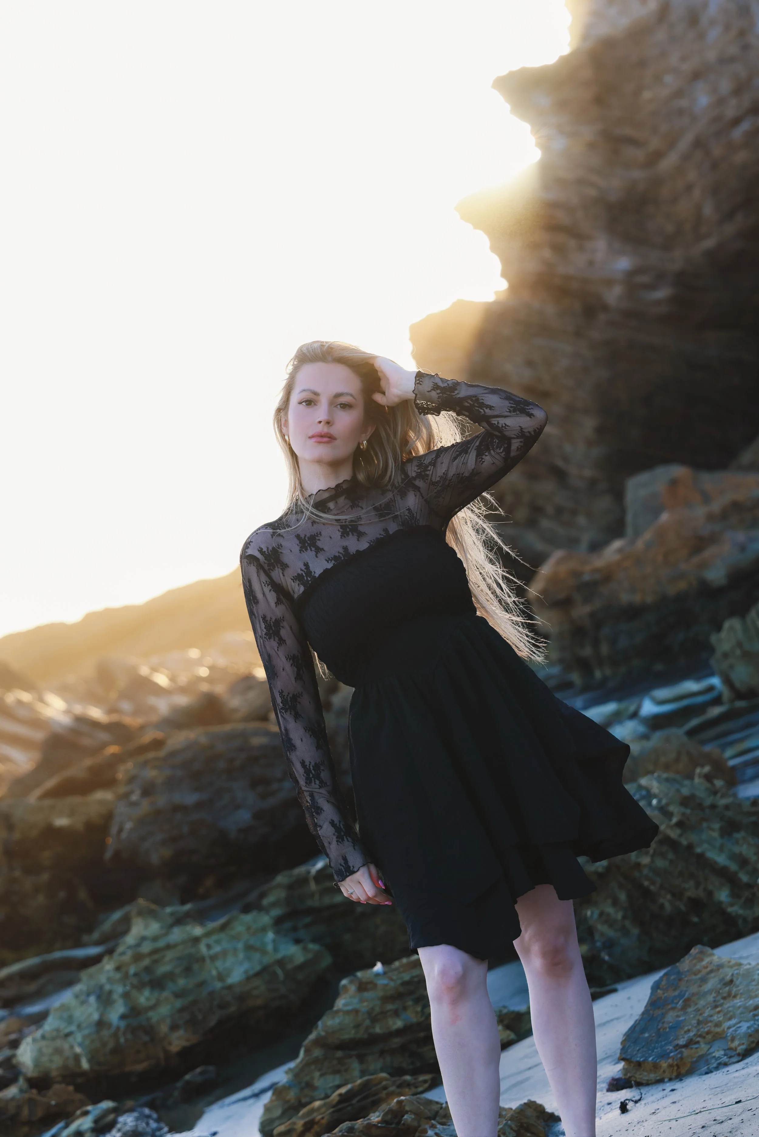 A woman in a black lace dress standing on rocky beach during sunset, with rocks and ocean in the background.